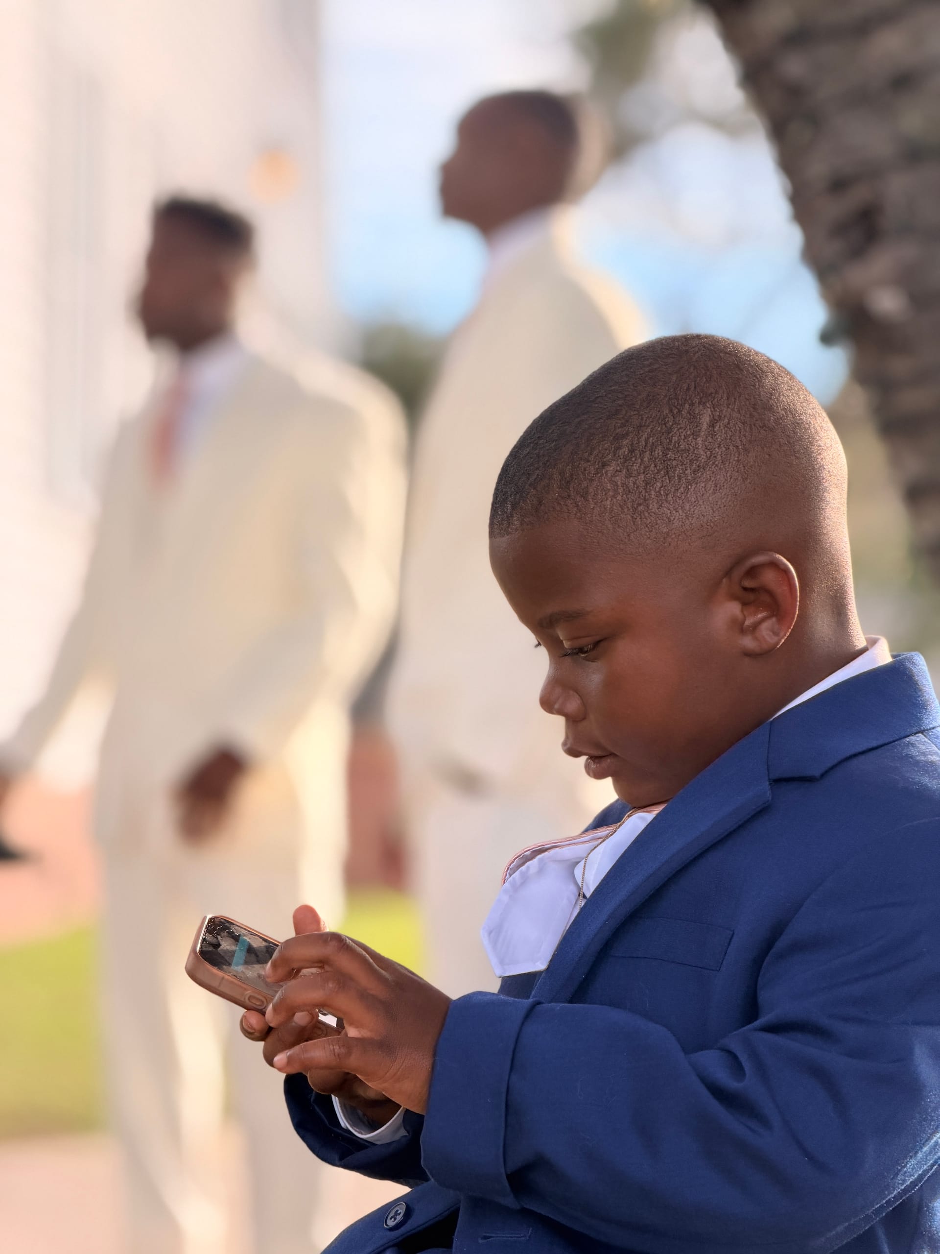 A young boy dressed in a navy suit looks down at a smartphone during an outdoor wedding ceremony while groomsmen in cream suits stand blurred in the background.