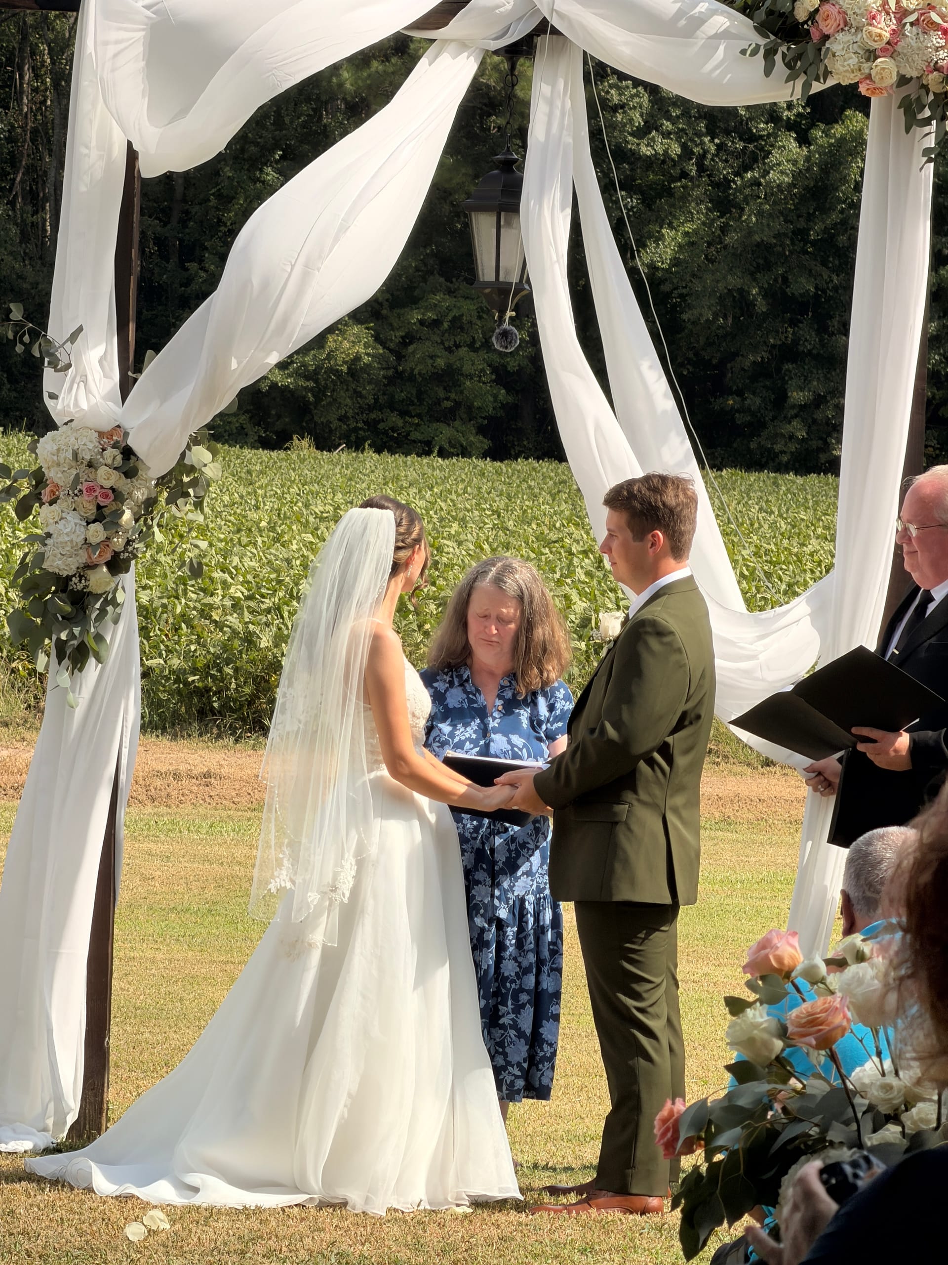 Bride and groom hold hands during their outdoor wedding ceremony beneath a white-draped wooden arch adorned with blush florals, with lush farmland fields stretching behind them at Scarborough House.