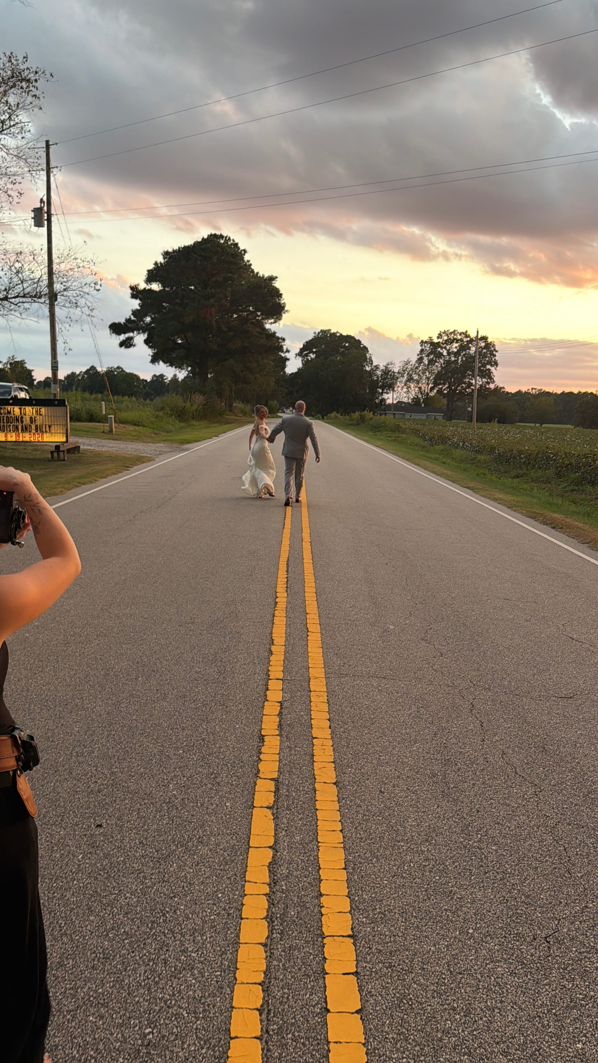 Bride and groom walk hand in hand down a two-lane road at sunset near Scarborough House wedding venue, with warm golden skies and lush trees framing the scene.