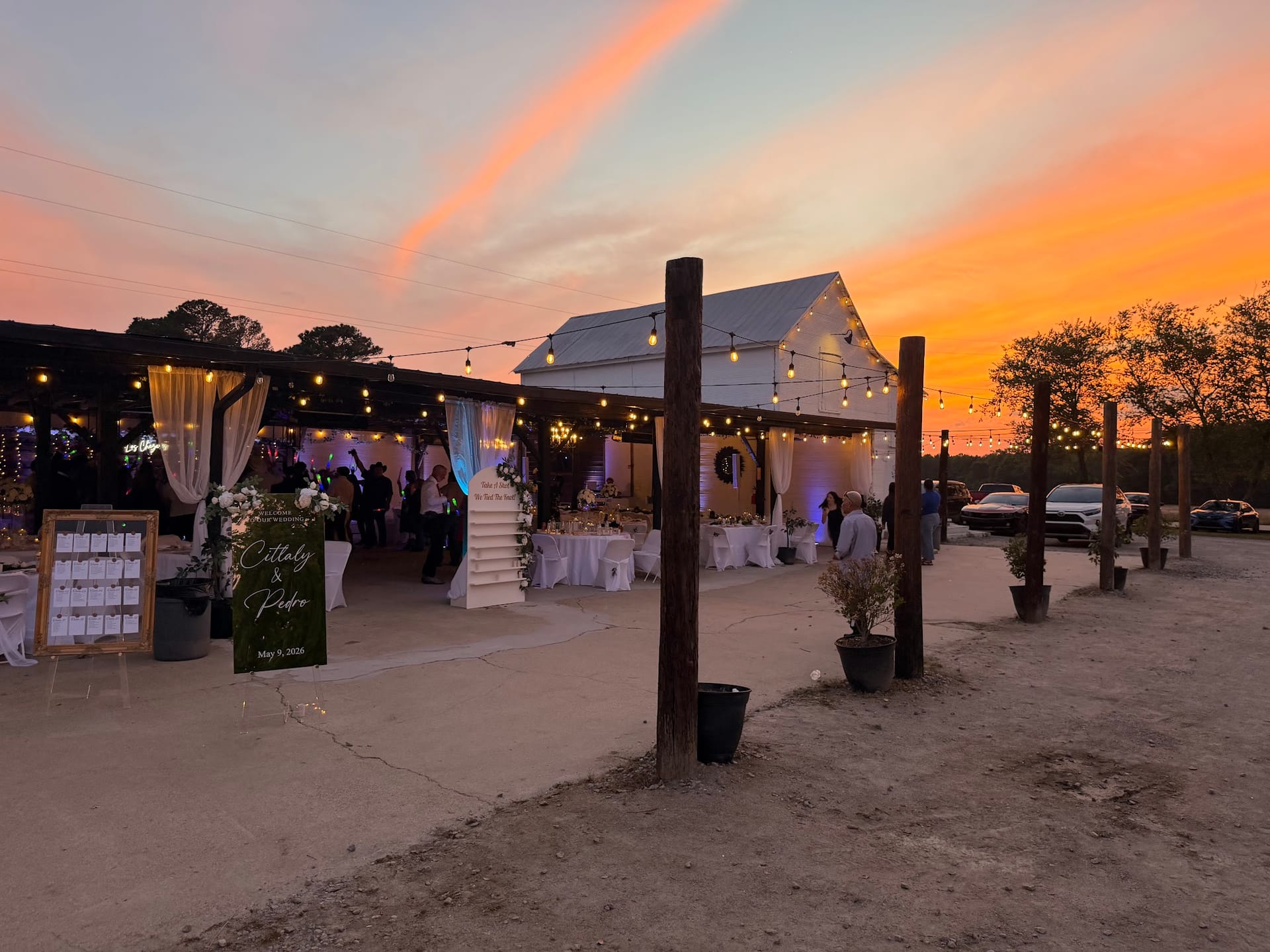 Outdoor wedding reception at dusk with glowing string lights over a white tent venue, a vibrant orange and pink sunset sky, and a green seating chart sign for Cittaly and Pedro dated May 9 2026.