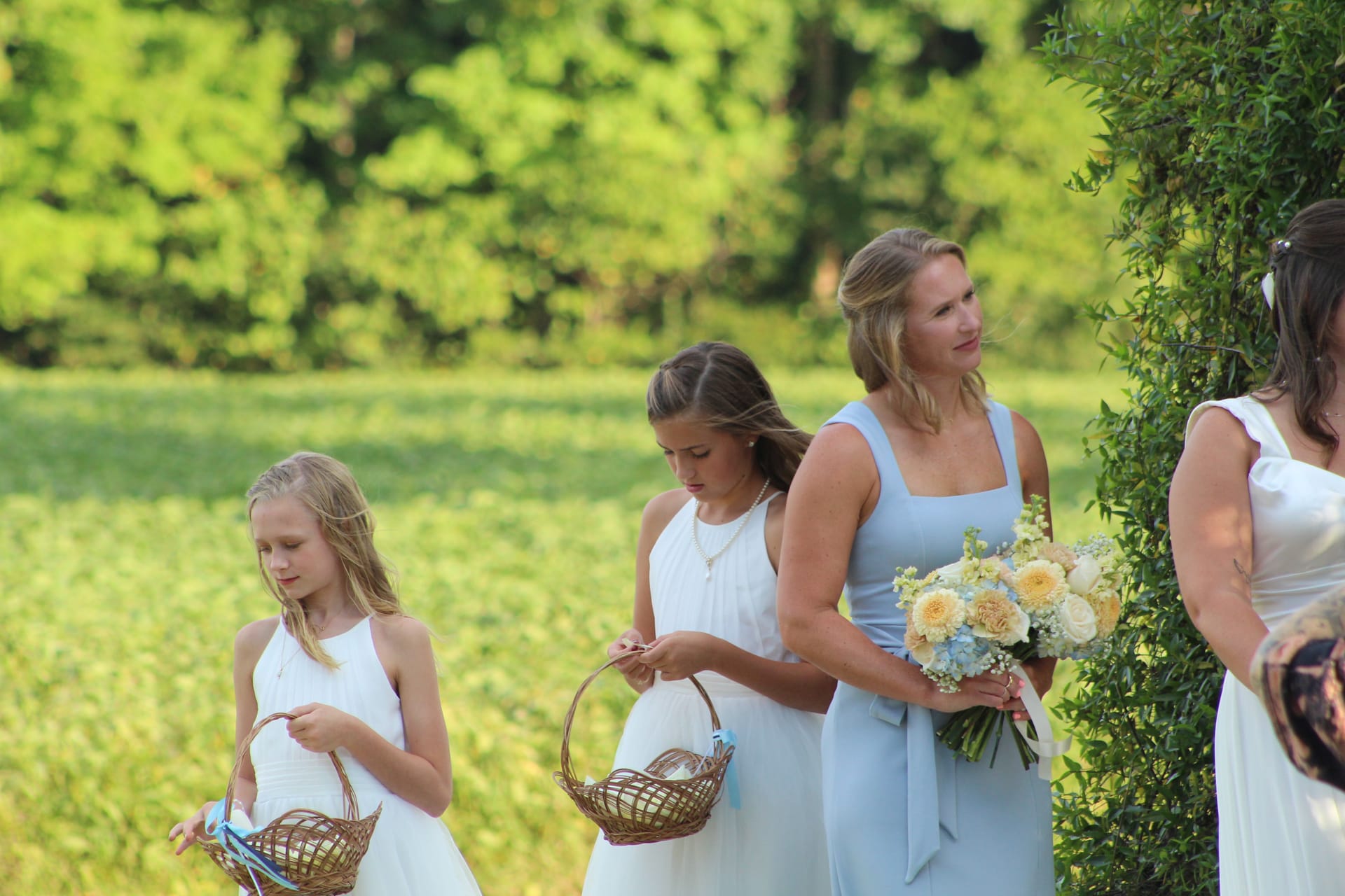 Two flower girls in white dresses carrying wicker baskets walk alongside a bridesmaid in a light blue gown holding a pastel bouquet during an outdoor wedding ceremony at a historic estate.