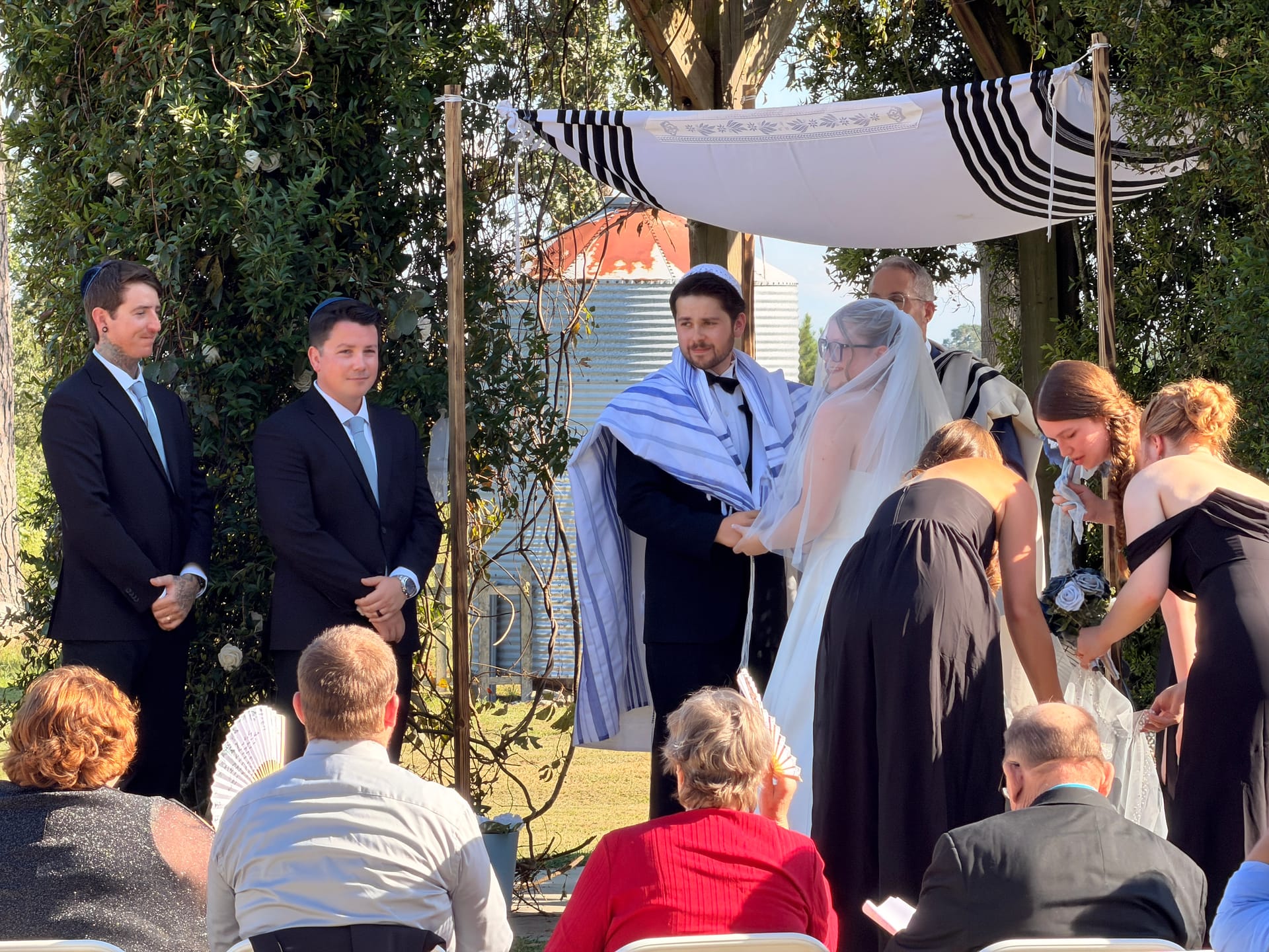 Bride and groom exchange vows beneath a tallit chuppah on the ceremony lawn at Scarborough House, surrounded by bridesmaids and groomsmen with lush greenery and farmland in the background.