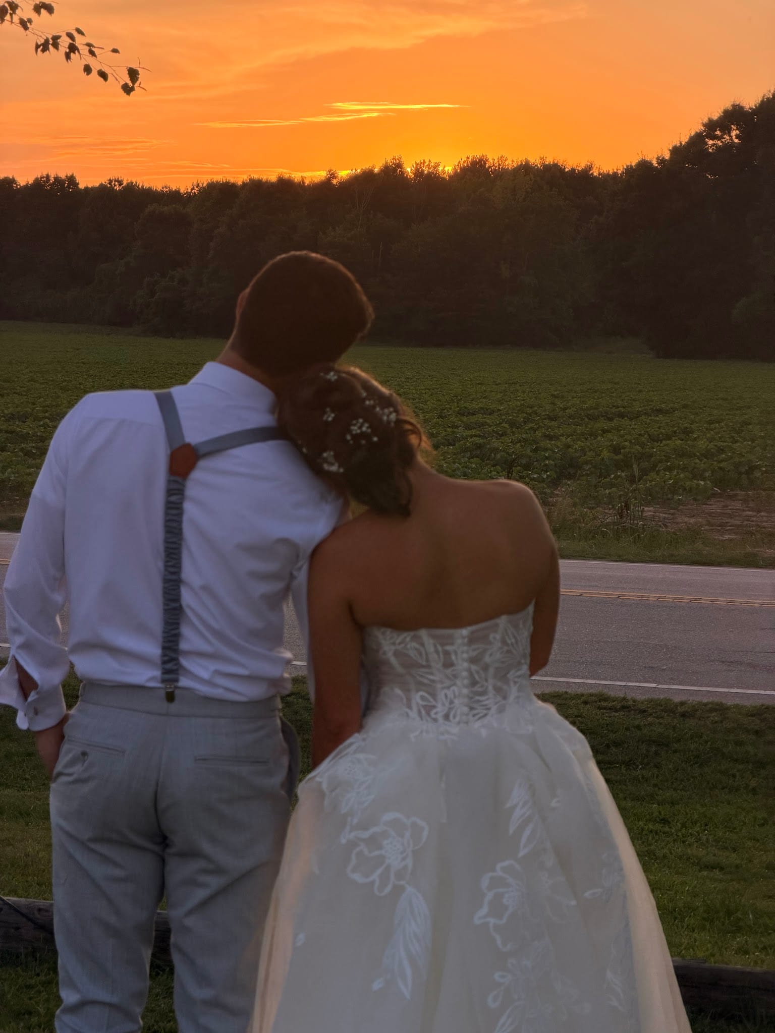 Bride and groom standing together from behind gazing at a vivid orange sunset over open farmland fields at Scarborough House wedding venue.