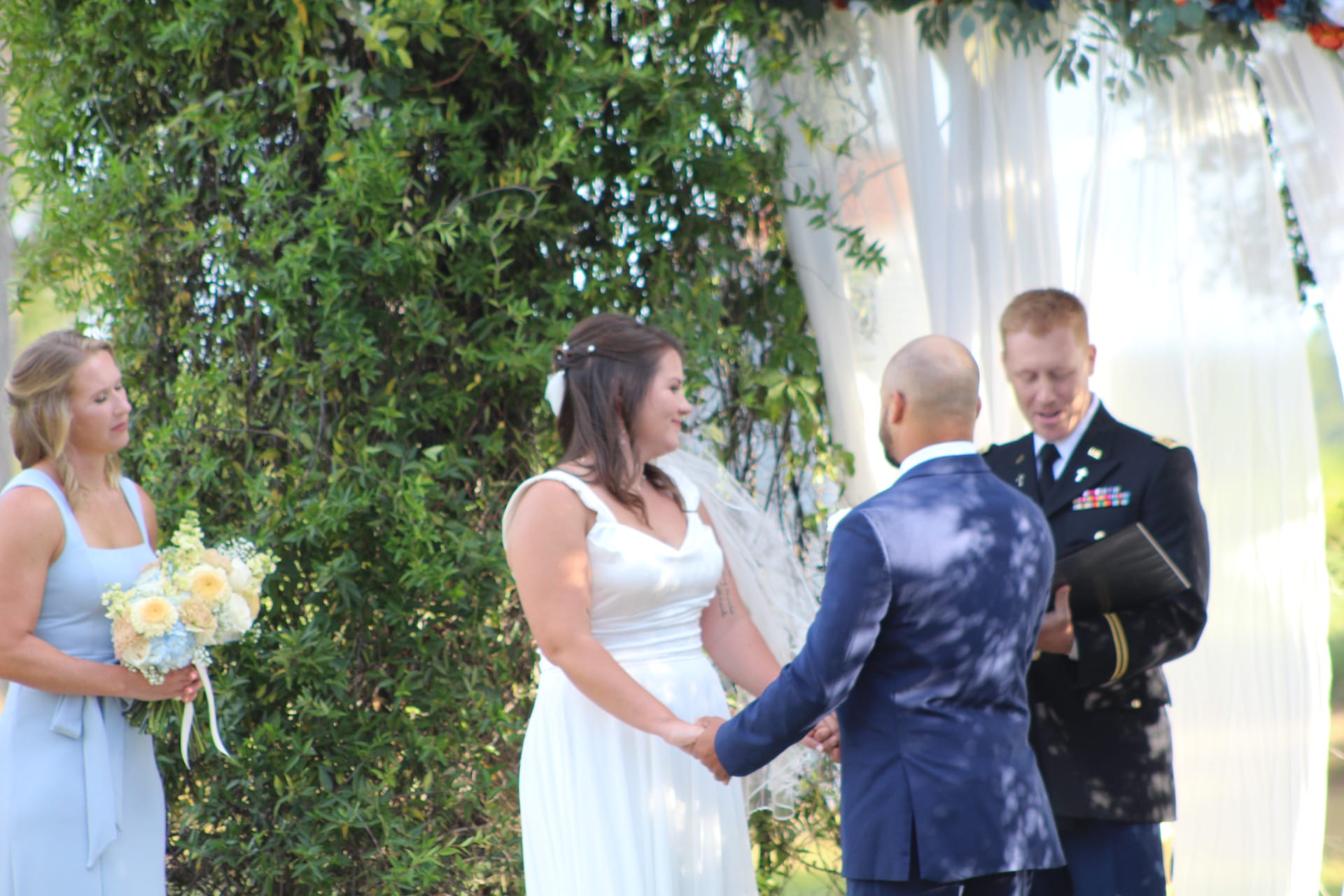 Bride in white gown holds hands with groom during outdoor wedding ceremony officiated by a military officer beside a white draped arch and lush green trees.