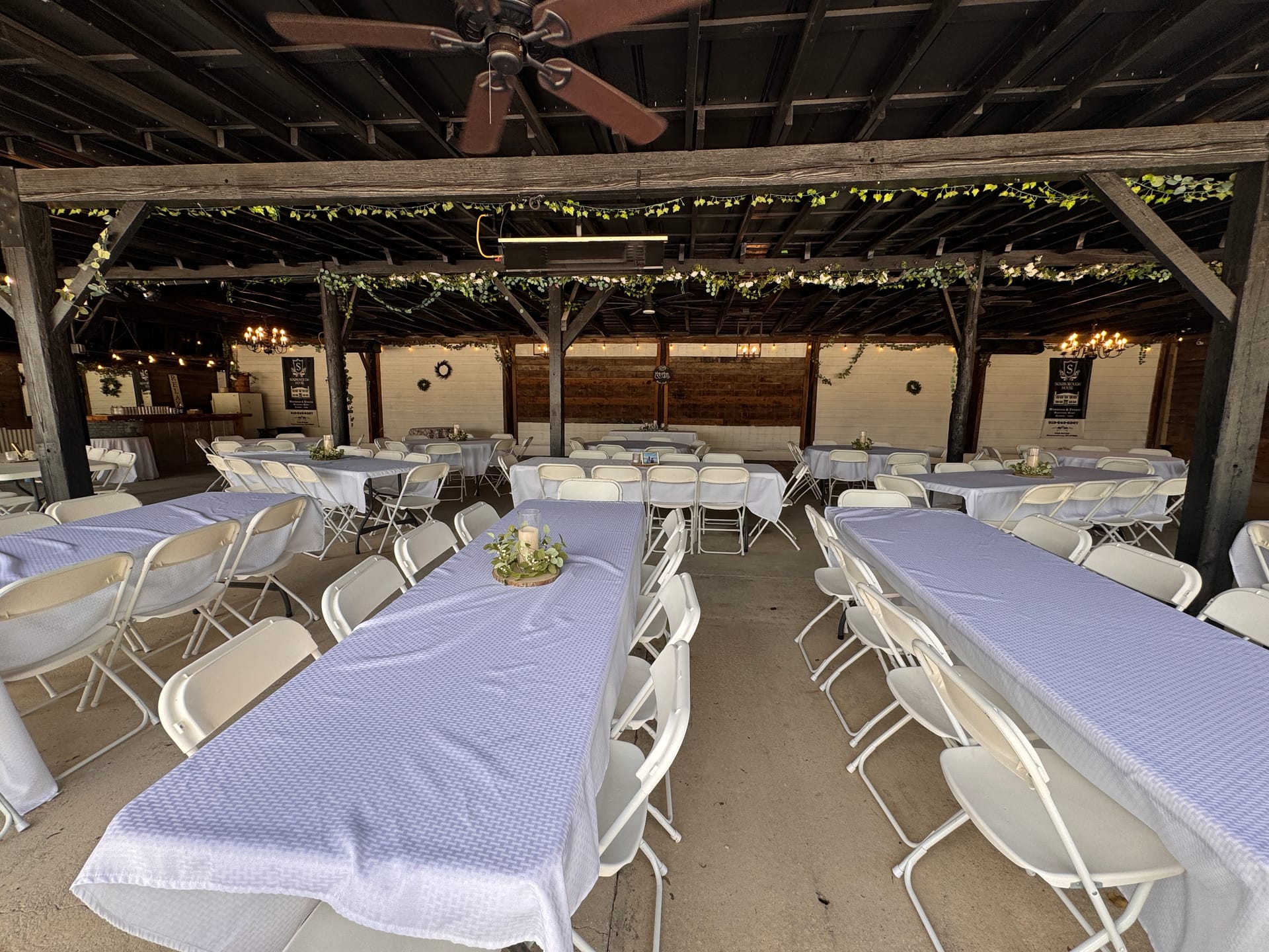 Rustic open-air pavilion at Scarborough House set for a wedding reception with white folding chairs, blue-checked tablecloths, greenery garland draped across wooden beams, and warm chandelier lighting.