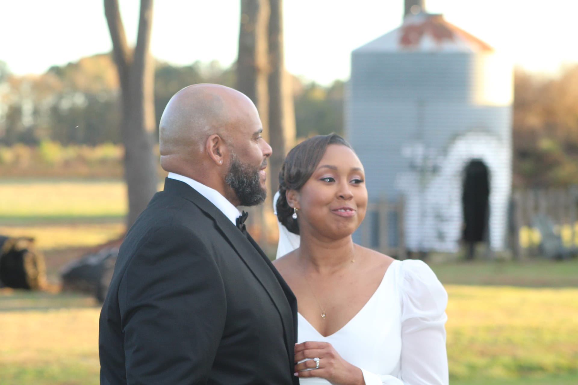 Bride in a white long-sleeve gown smiles warmly while holding the arm of her tuxedo-clad groom during a golden-hour portrait on the open farmland grounds of Scarborough House historic estate.
