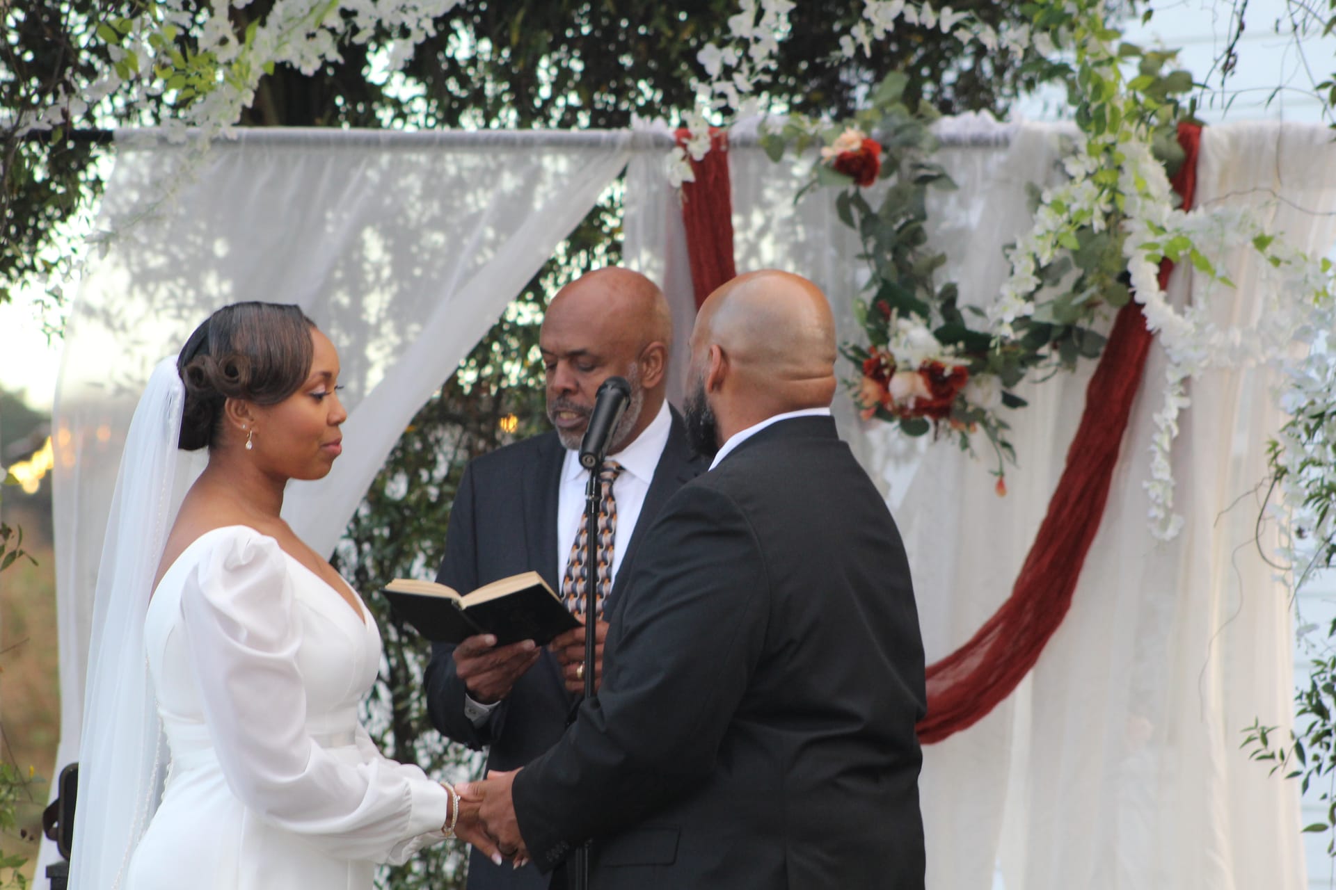 Bride in a white off-shoulder gown holds hands with her groom as an officiant reads from a book beneath a draped floral arch adorned with white blooms and burgundy accents at an outdoor wedding ceremony.
