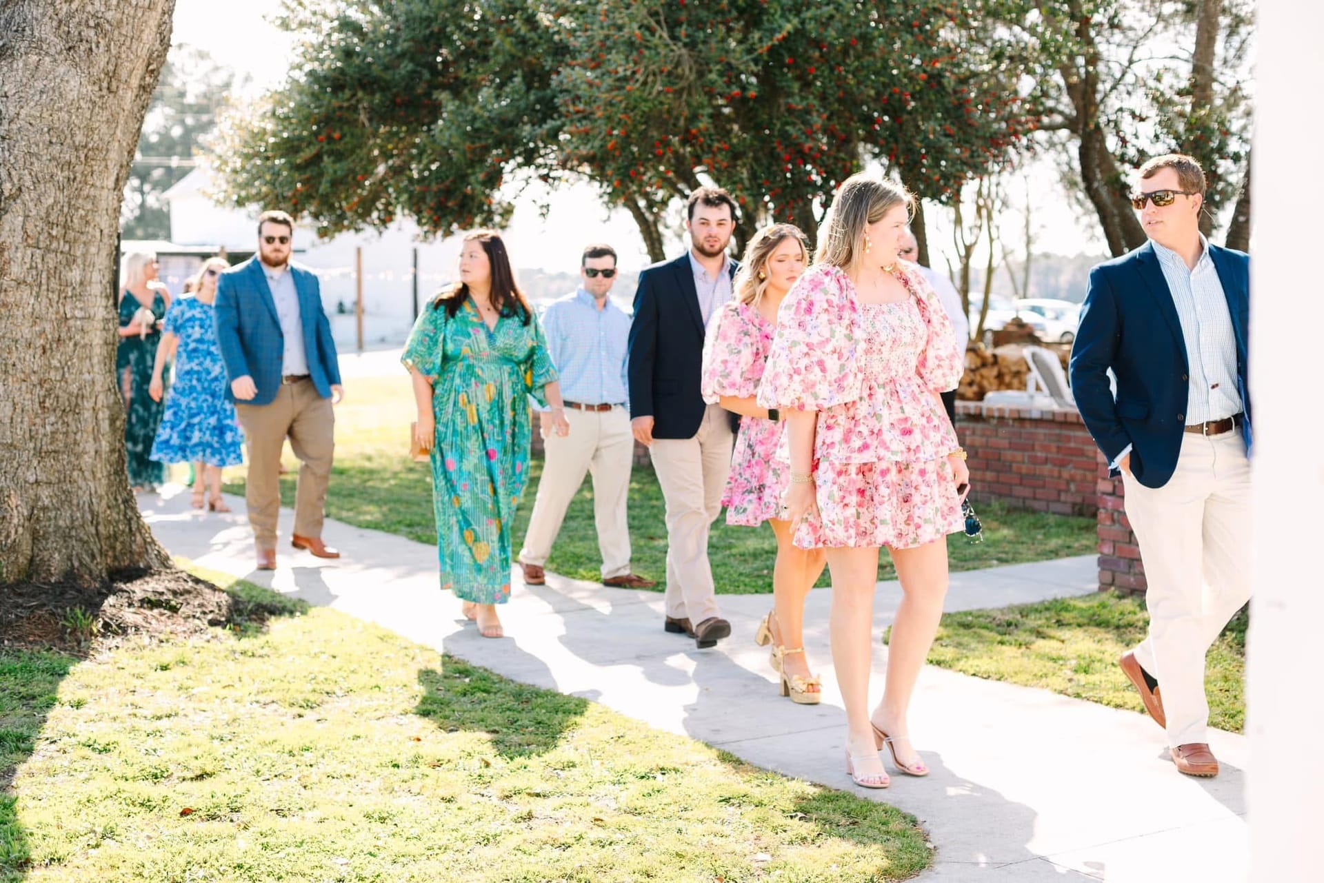 Elegantly dressed wedding guests in floral dresses and navy blazers stroll along a sunlit pathway beneath oak trees at Scarborough House historic estate venue.