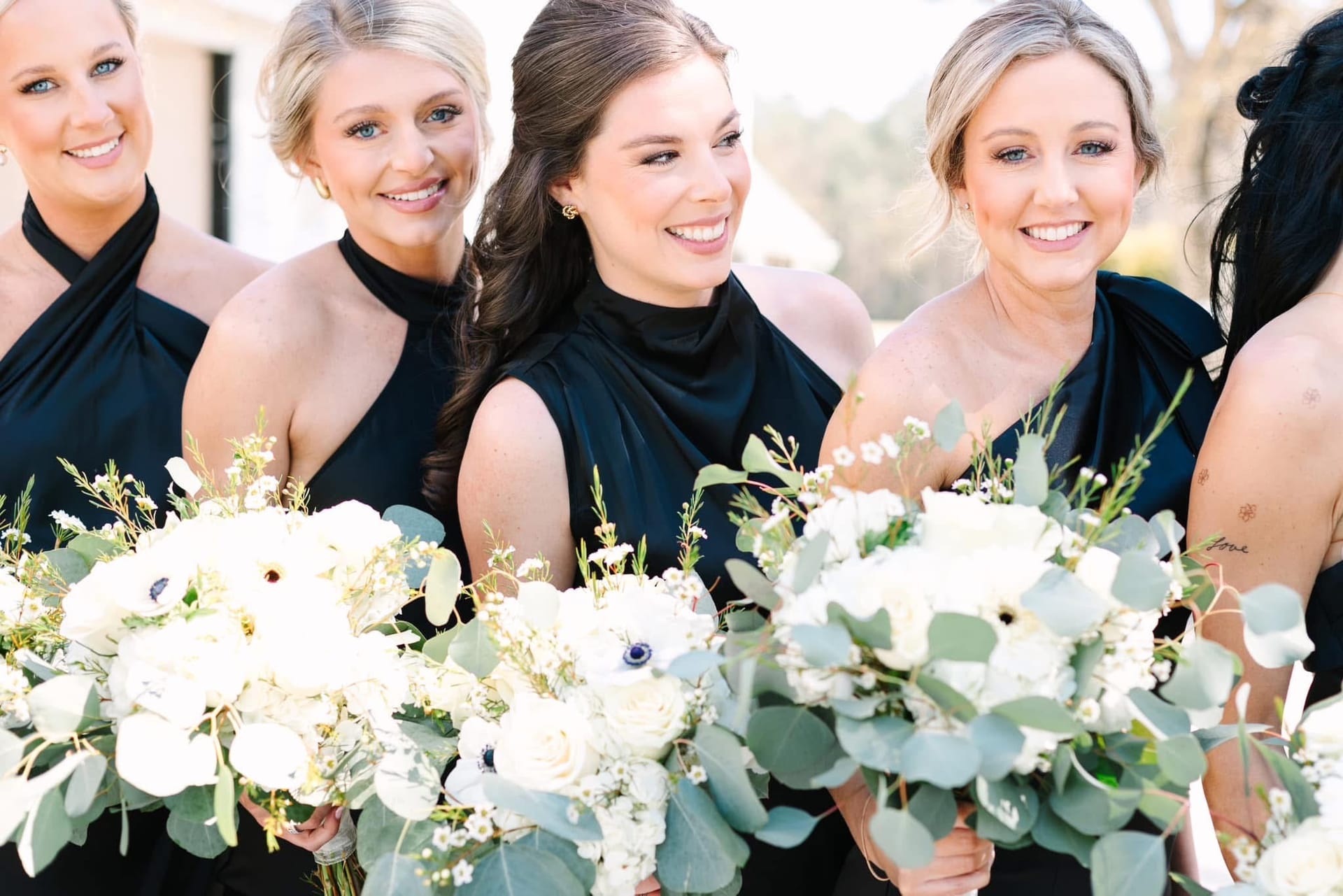 Four bridesmaids in elegant black halter gowns laughing and smiling together while holding lush white floral and eucalyptus bouquets at Scarborough House.