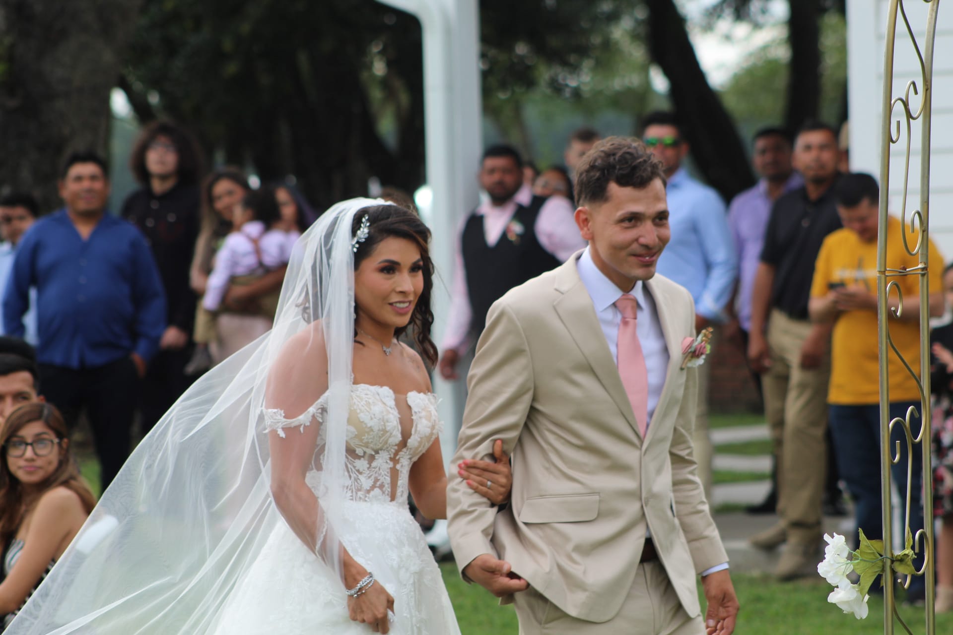 Bride in a lace ball gown and groom in a tan suit with pink tie walk arm in arm down the outdoor ceremony aisle at Scarborough House surrounded by smiling guests.