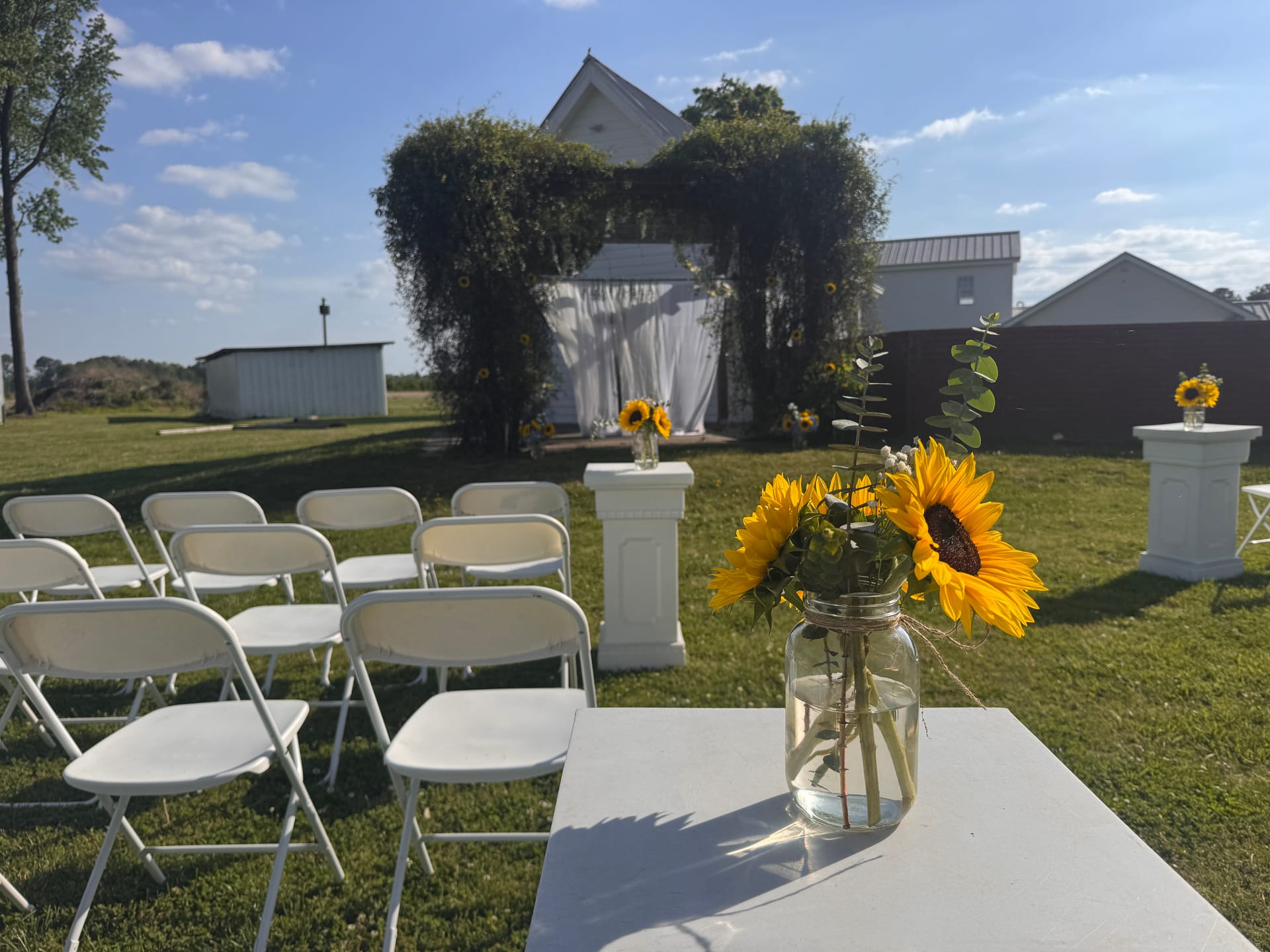 Sunflowers in a mason jar vase on a white pedestal overlooking rows of white folding chairs and an ivy-covered wedding ceremony arch on the ceremony lawn at Scarborough House.
