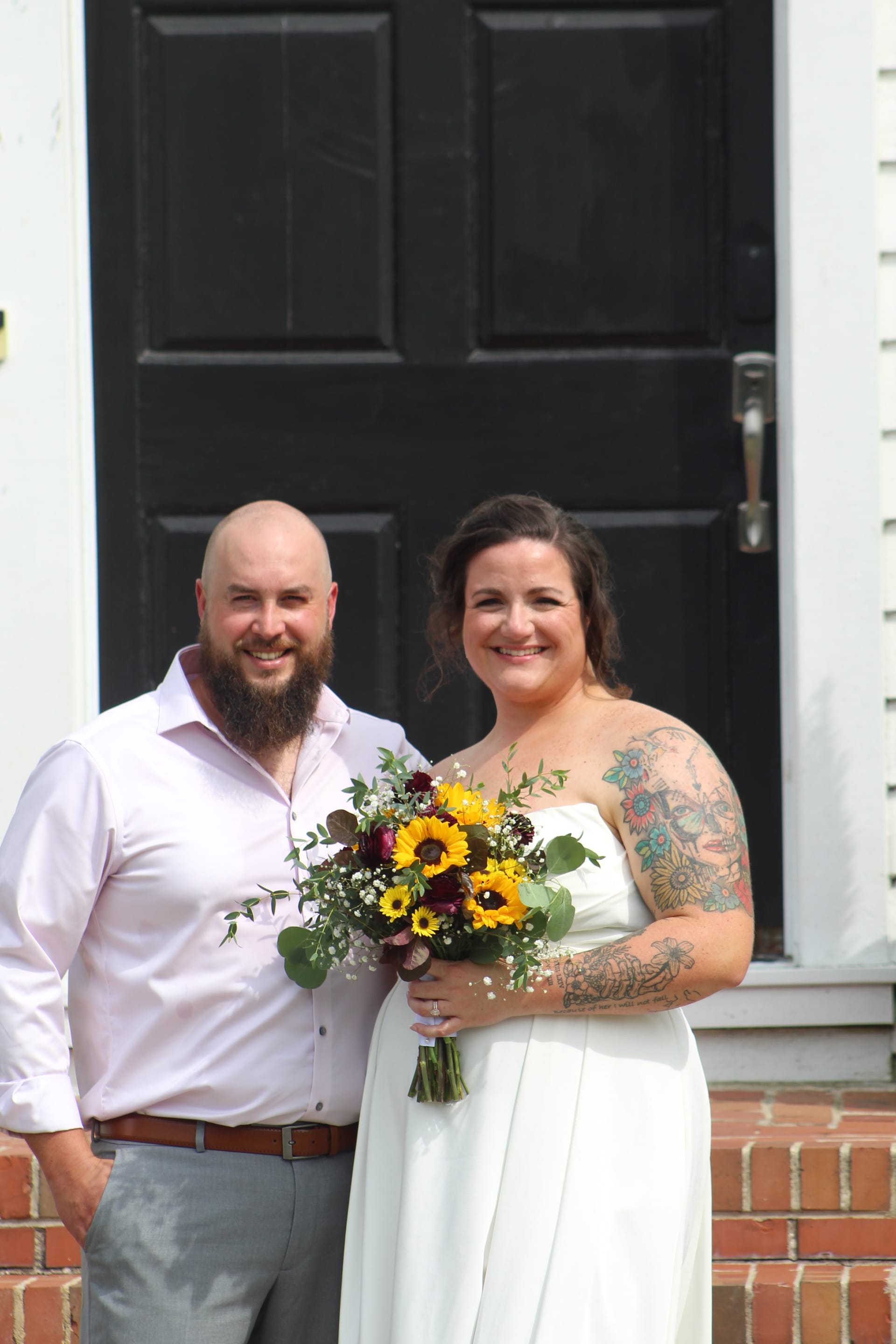 Smiling bride in a white strapless gown holding a sunflower and burgundy bouquet stands beside her bearded groom in a pink shirt in front of the historic black door at Scarborough House.