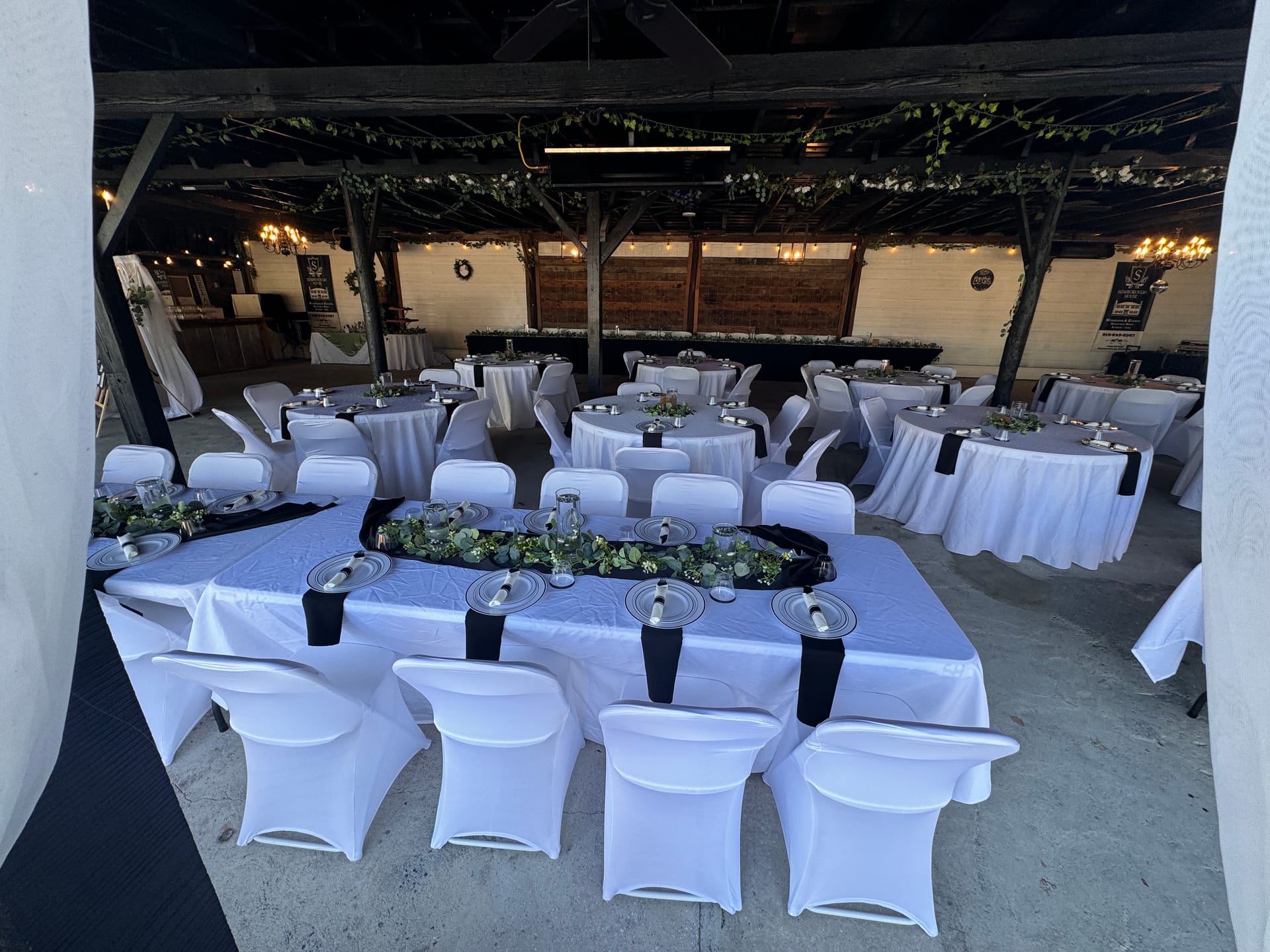 Elegantly set reception hall at Scarborough House featuring white linen tables with black napkins, silver chargers, and lush greenery runners beneath a rustic wooden ceiling draped with ivy and warm string lights.