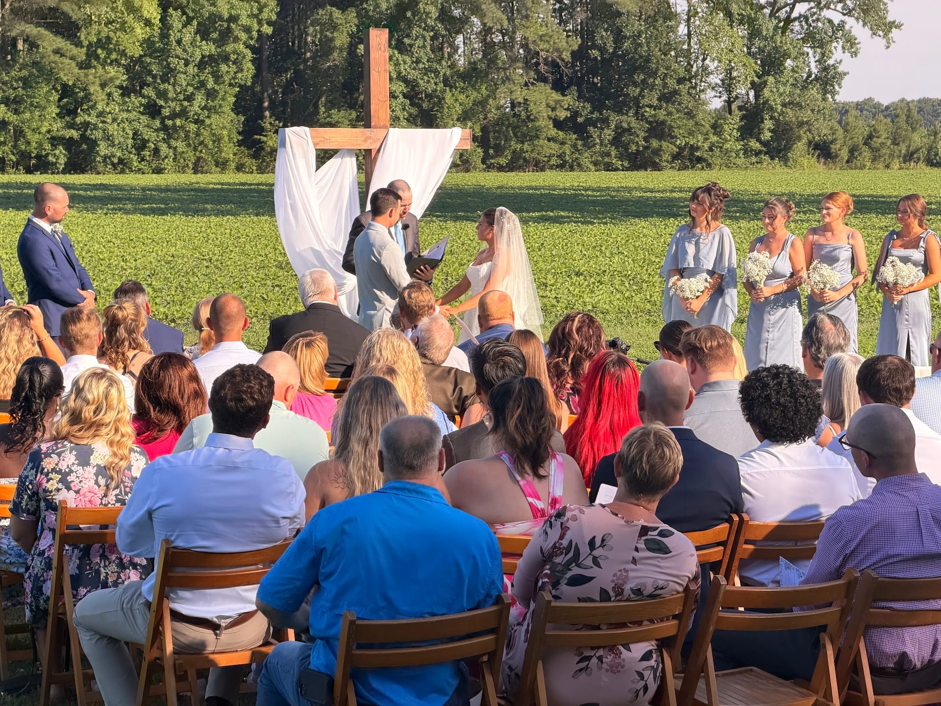 Outdoor wedding ceremony at a historic estate venue with guests seated facing a couple exchanging vows before a large wooden cross draped in white fabric, flanked by bridesmaids in blue against a backdrop of green farmland fields and trees.