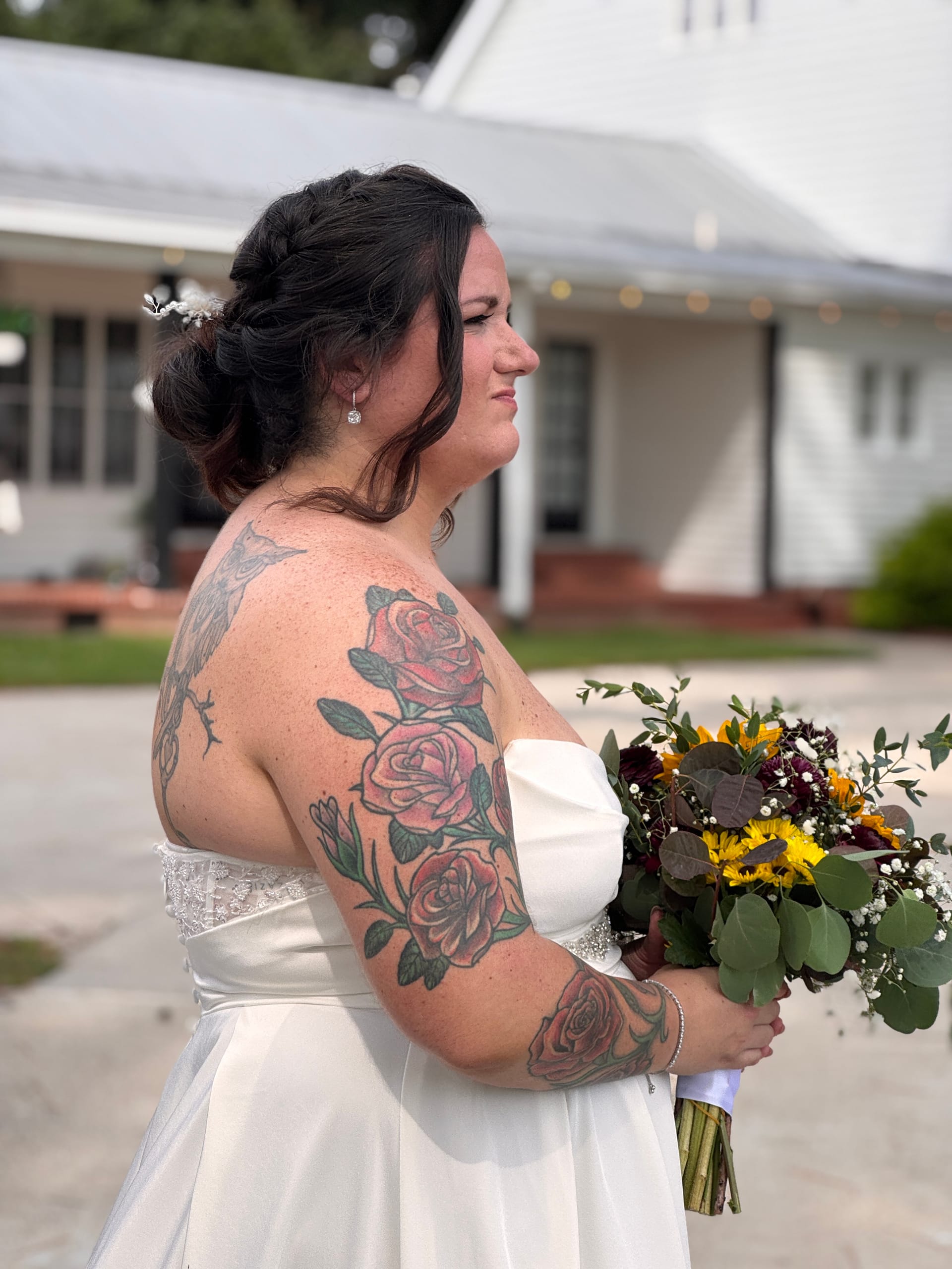 A bride in a strapless white gown with an elaborate rose tattoo sleeve holds a sunflower and burgundy bouquet in front of the white historic Scarborough House manor.
