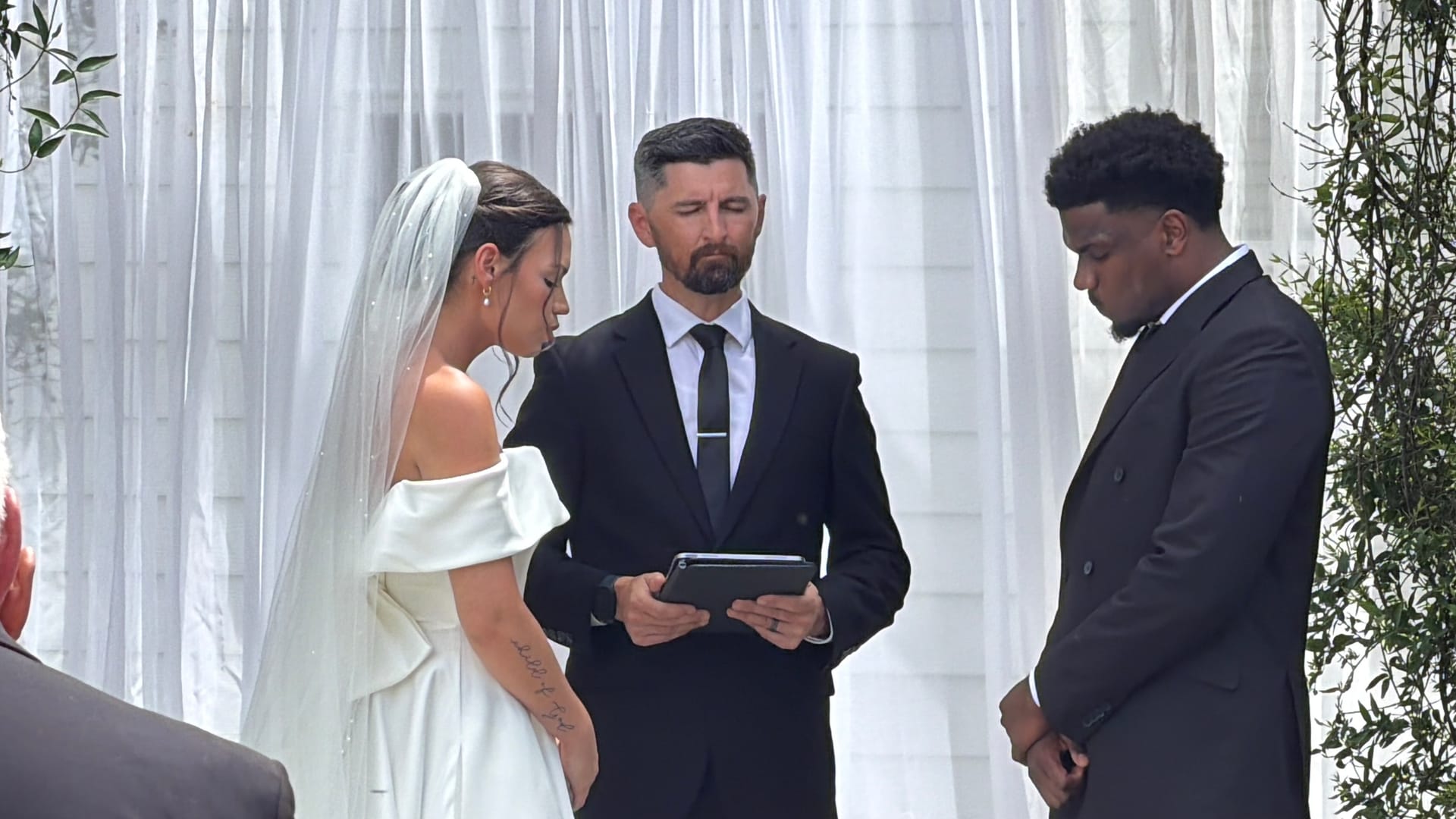 Bride in an off-shoulder white gown and veil stands with the groom in a dark suit as an officiant reads from a tablet during an outdoor wedding ceremony against a draped white backdrop.