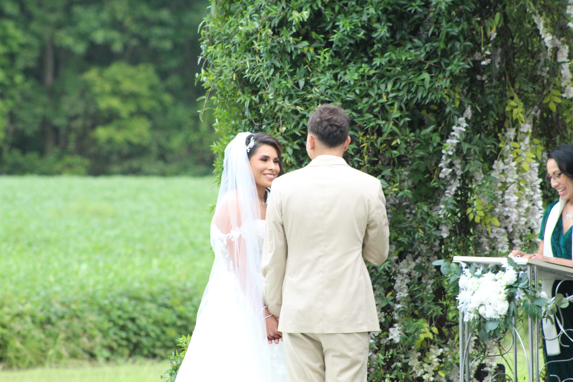 Smiling bride in white gown and veil holds hands with groom in tan suit during an outdoor wedding ceremony beneath a lush green and white floral arch at Scarborough House.