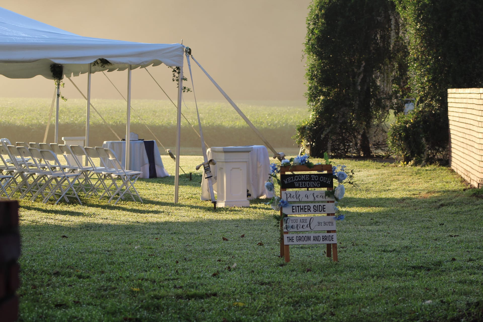 Outdoor wedding ceremony lawn at Scarborough House featuring white folding chairs under a frame tent and a rustic wooden welcome sign decorated with blue flowers in warm golden-hour light.