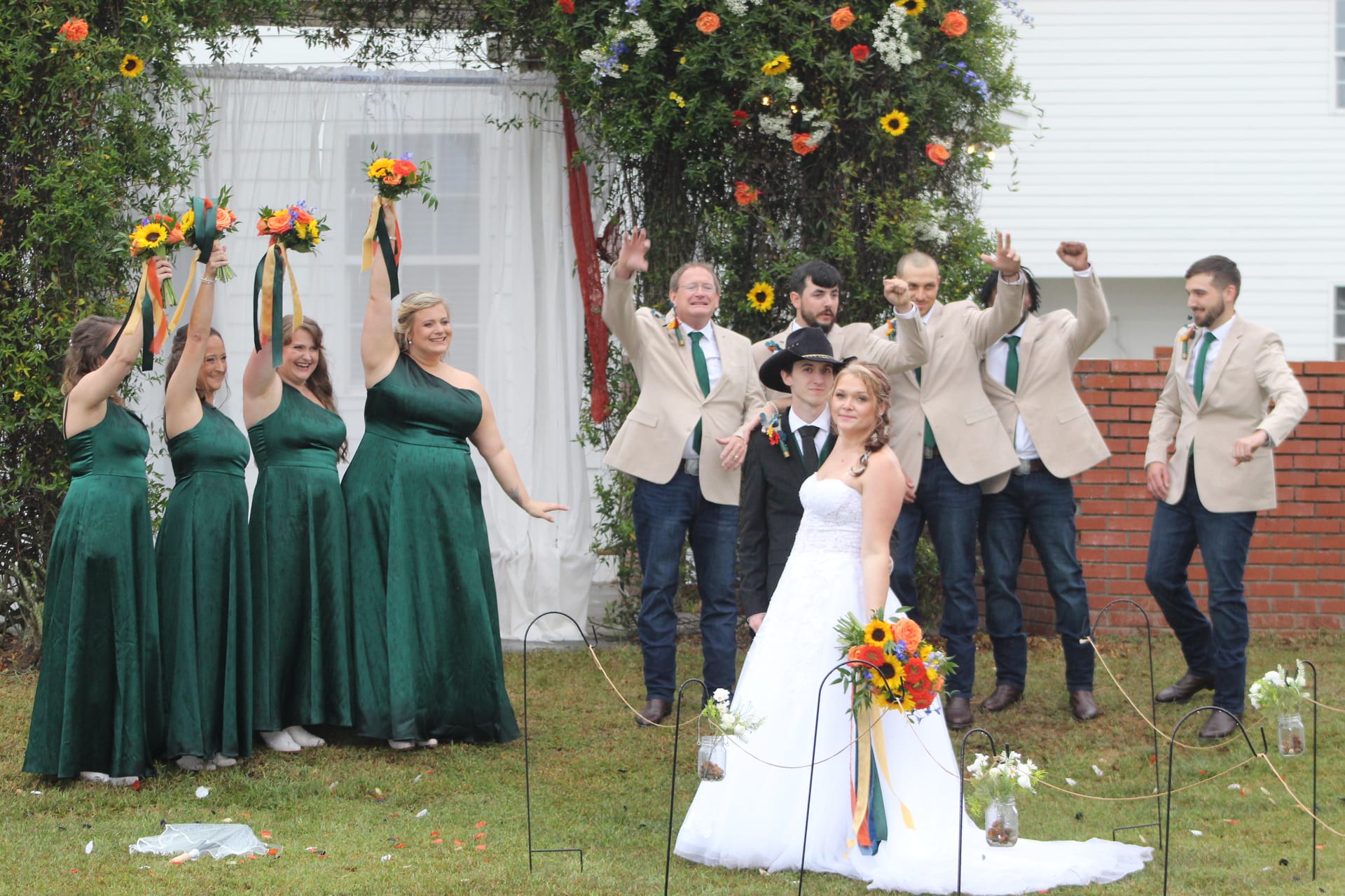 Bride holding a sunflower and wildflower bouquet poses in front of her celebrating wedding party beneath a floral arch on the ceremony lawn at Scarborough House historic estate.