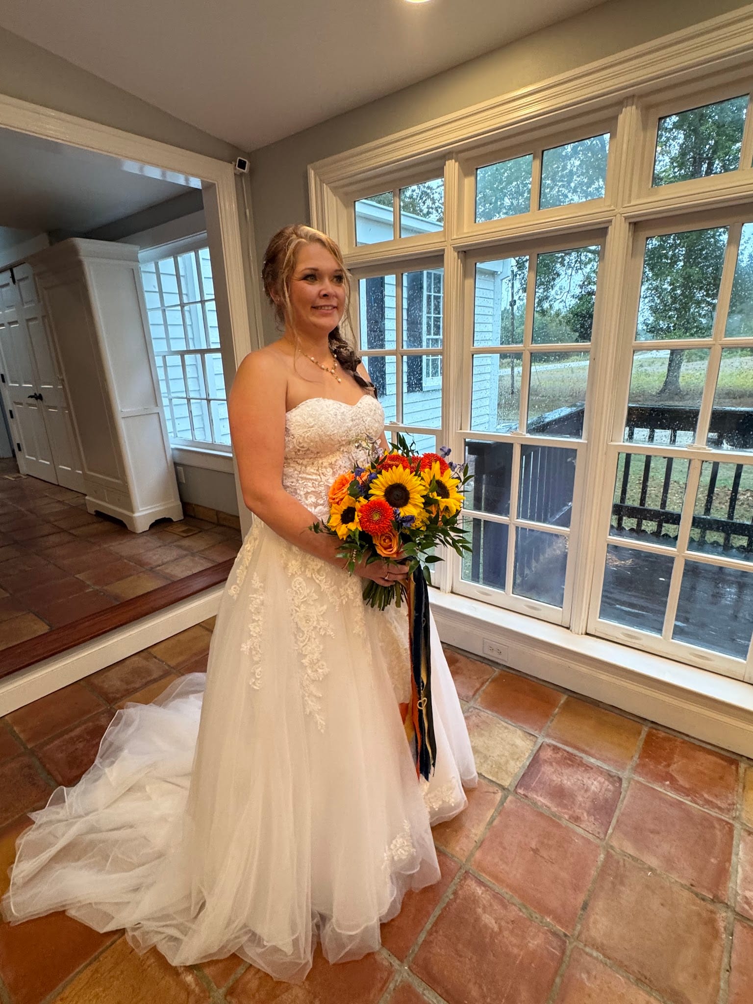 Bride in a strapless lace A-line wedding gown holding a vibrant sunflower and orange bouquet standing before large multi-pane windows at Scarborough House historic estate.