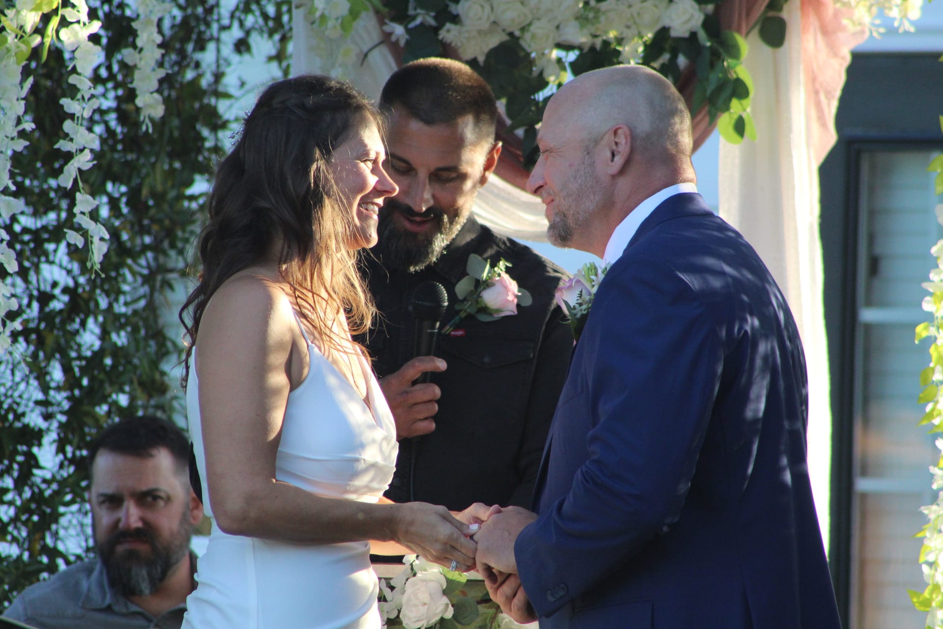 Bride in a white gown and groom in a navy suit share a joyful moment during their wedding vows beneath a floral arch at Scarborough House.