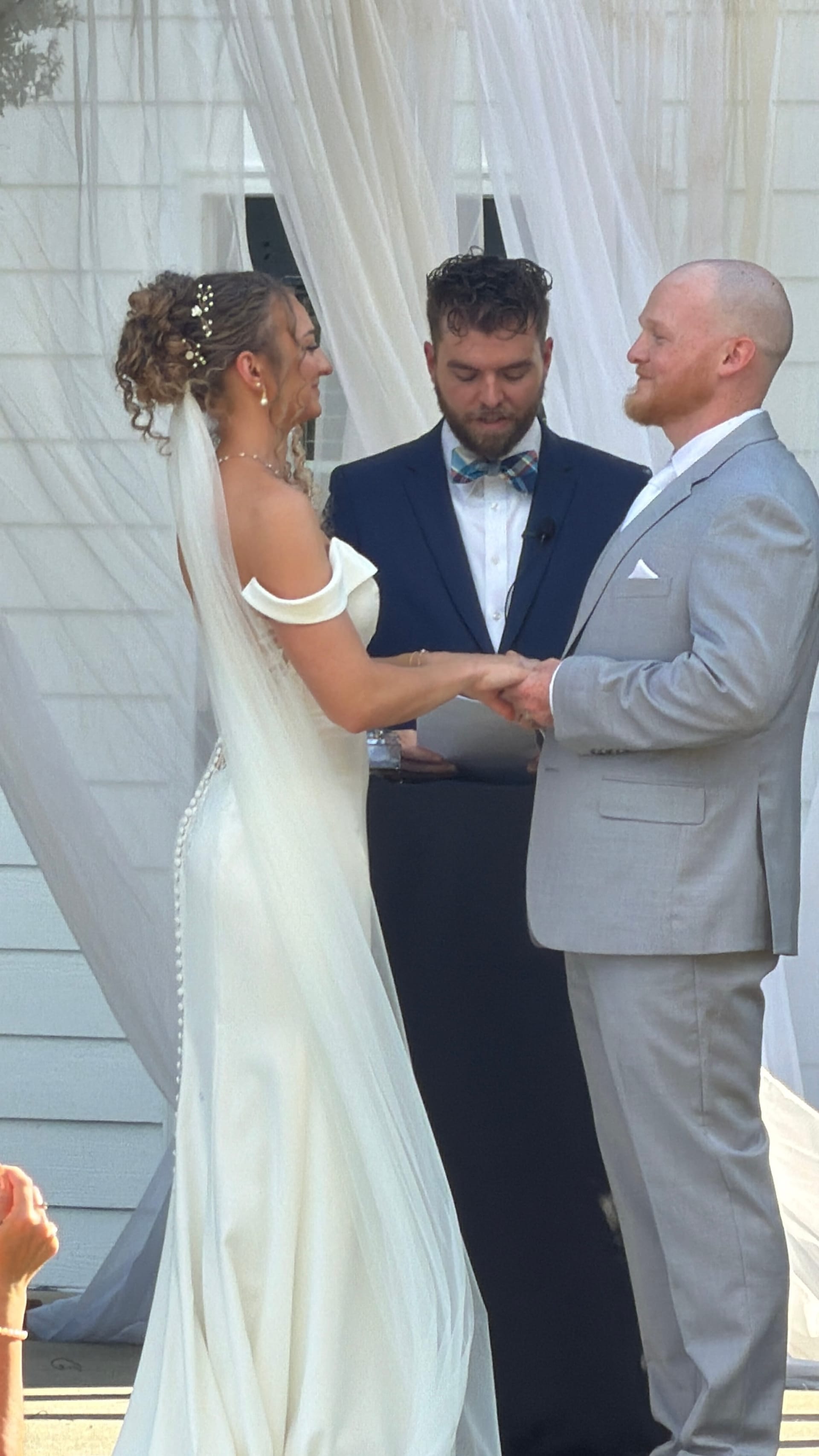 Bride and groom exchange vows and hold hands during an outdoor wedding ceremony at Scarborough House, with a navy-suited officiant reading from a booklet beneath flowing white drapery.