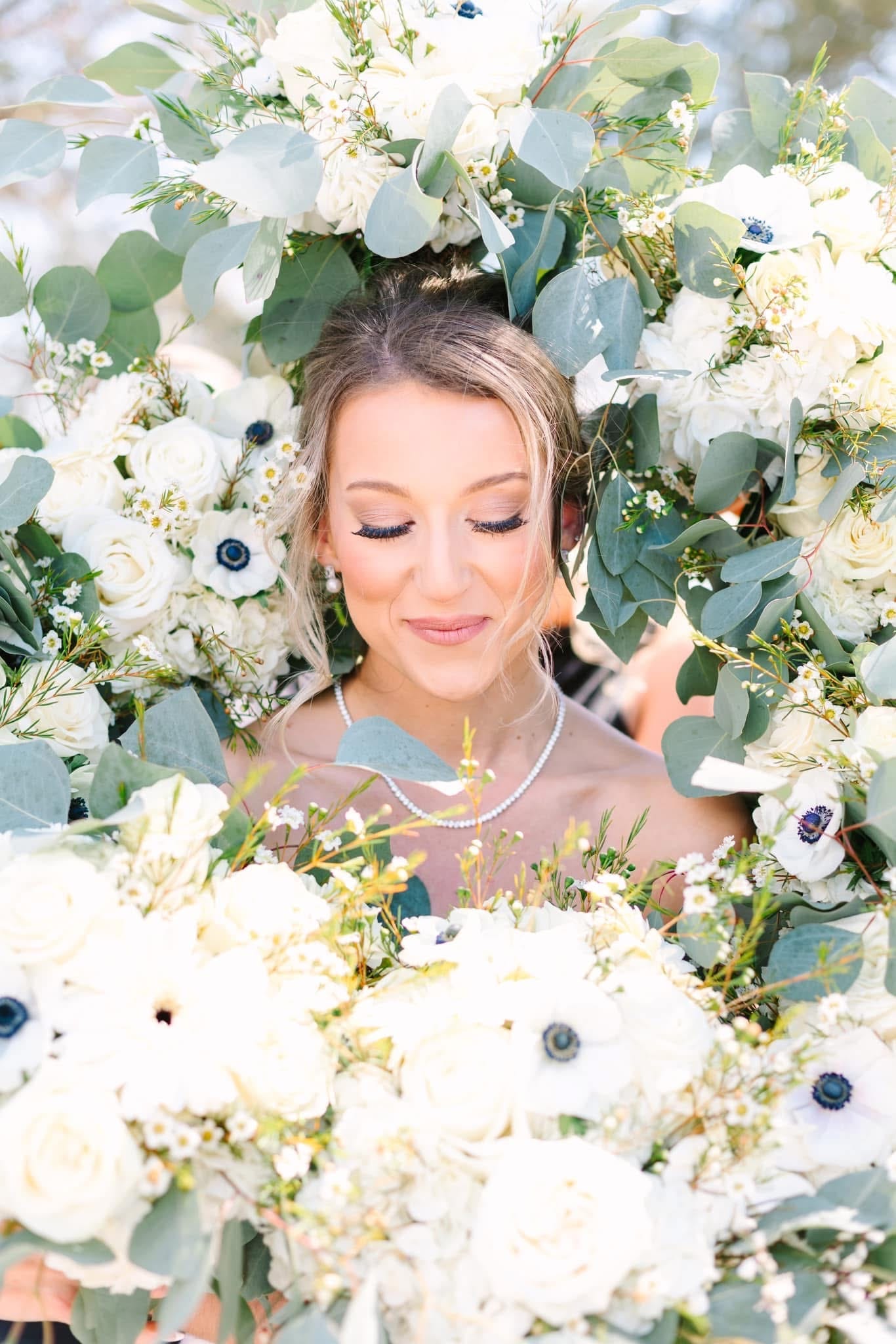 Bride with eyes softly closed smiles serenely while encircled by an abundant arrangement of white roses, anemones, and eucalyptus greenery at Scarborough House.
