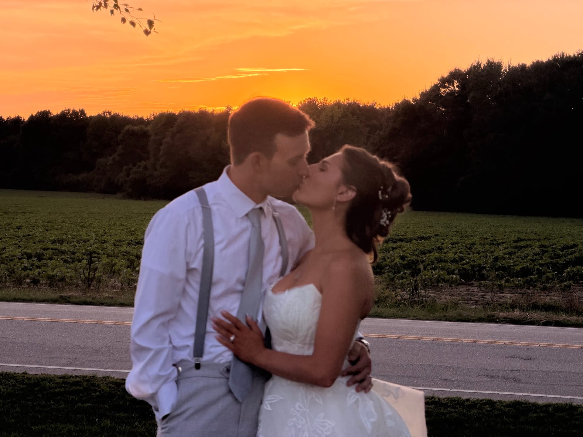 Bride in a strapless white gown and groom in a white shirt with gray suspenders share a kiss against a vivid orange sunset sky overlooking lush farmland fields at Scarborough House.