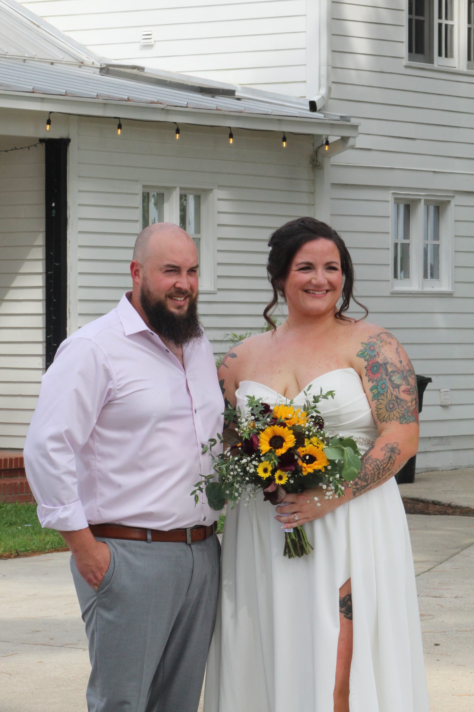 Smiling bride in a white strapless gown holding a sunflower bouquet stands beside the groom in a white dress shirt in front of the historic white clapboard exterior of Scarborough House wedding venue.
