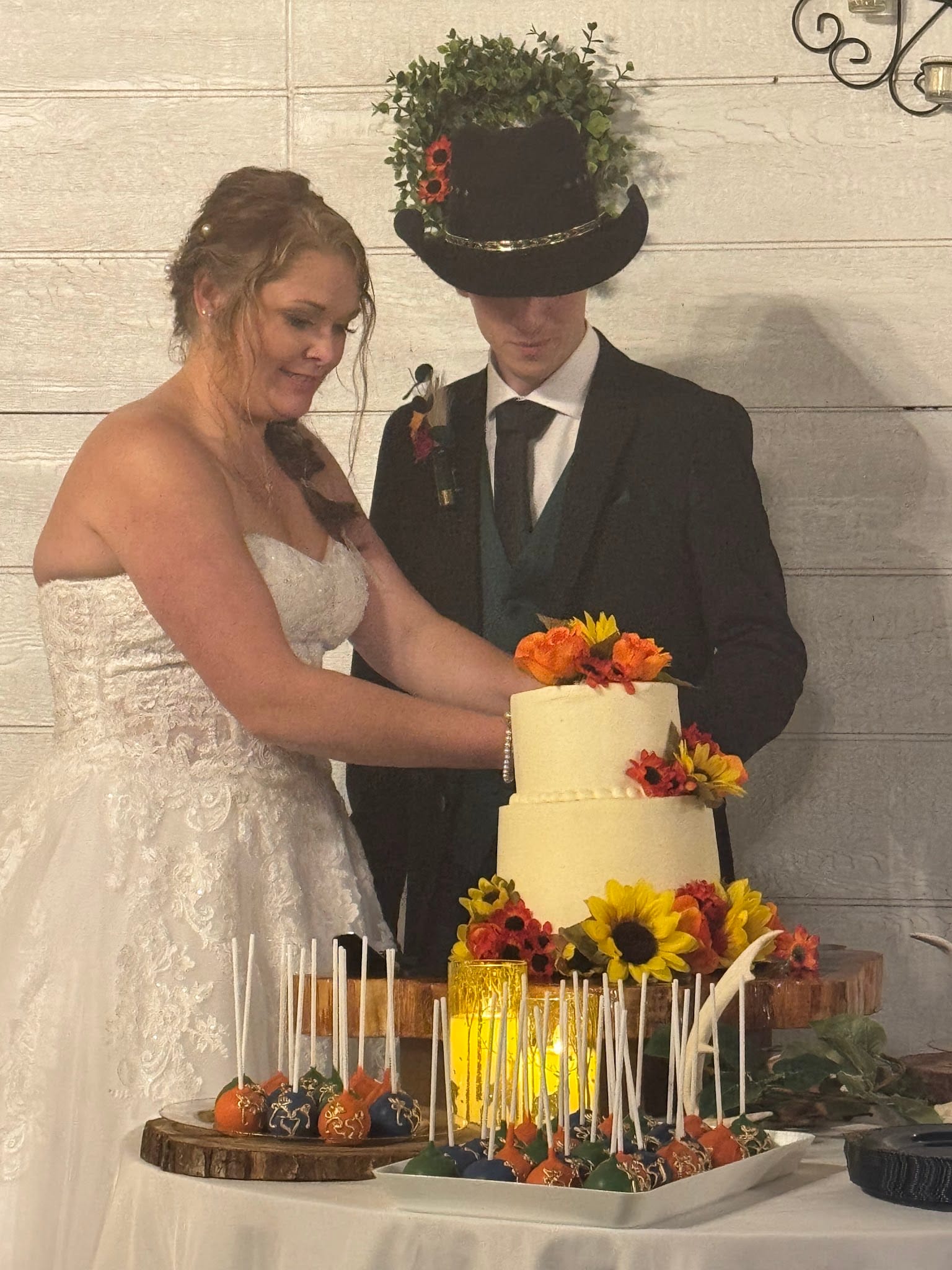 Bride in a lace strapless gown and groom in a dark suit and floral-adorned top hat cut their two-tier white wedding cake decorated with sunflowers and autumn blooms at Scarborough House.