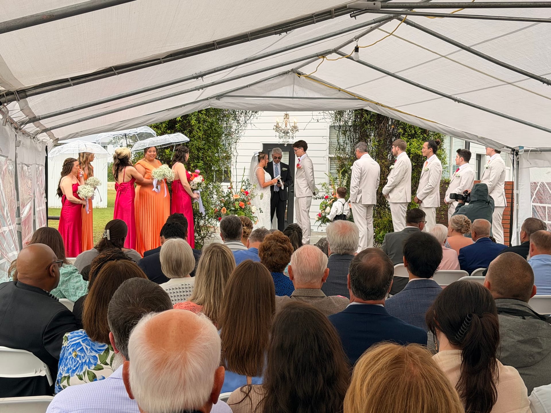 Wedding ceremony under a white marquee tent with colorful bridesmaids in orange and pink gowns standing opposite groomsmen in white suits as the couple exchanges vows before seated guests.