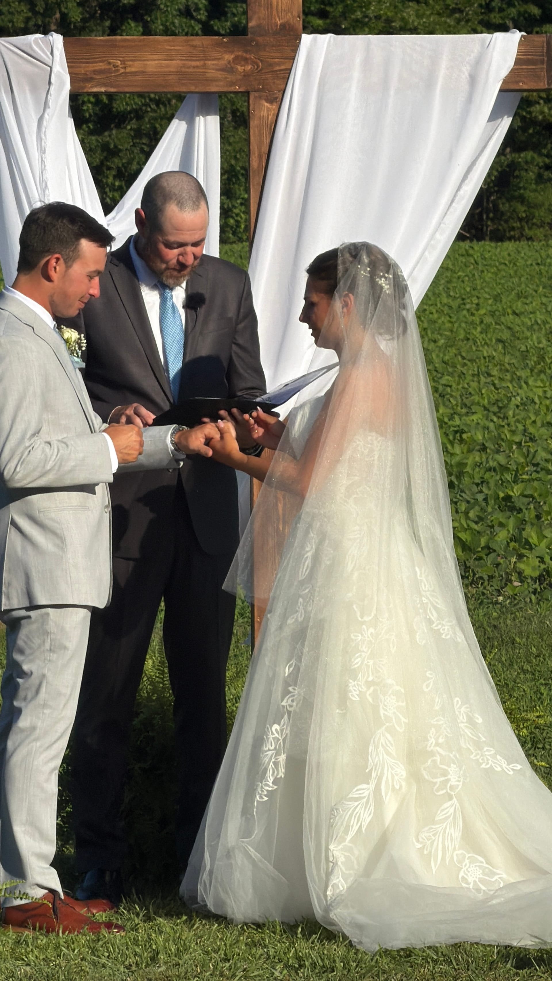 Bride and groom exchange rings during an outdoor wedding ceremony beneath a rustic wooden cross arbor draped with white fabric at Scarborough House estate.