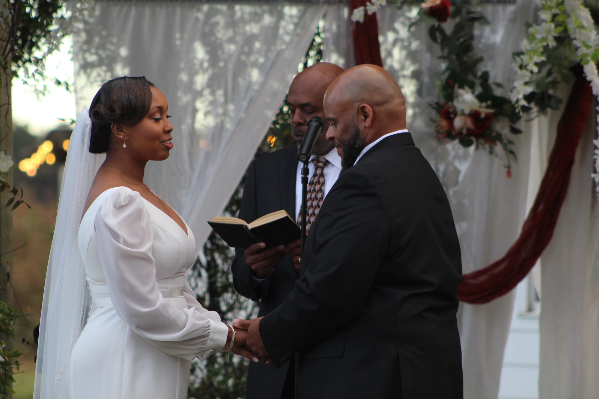 Bride and groom hold hands during outdoor wedding vow exchange beneath a white draped arch adorned with burgundy and white florals at Scarborough House.