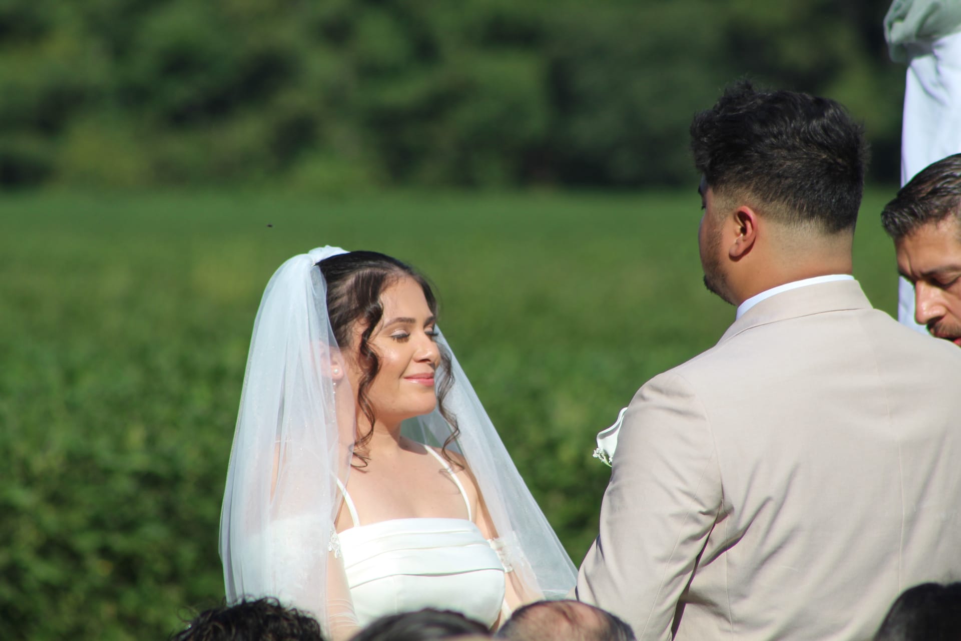 Bride in white gown and veil smiling softly at groom in a tan suit during an outdoor wedding ceremony at Scarborough House with lush green fields in the background.