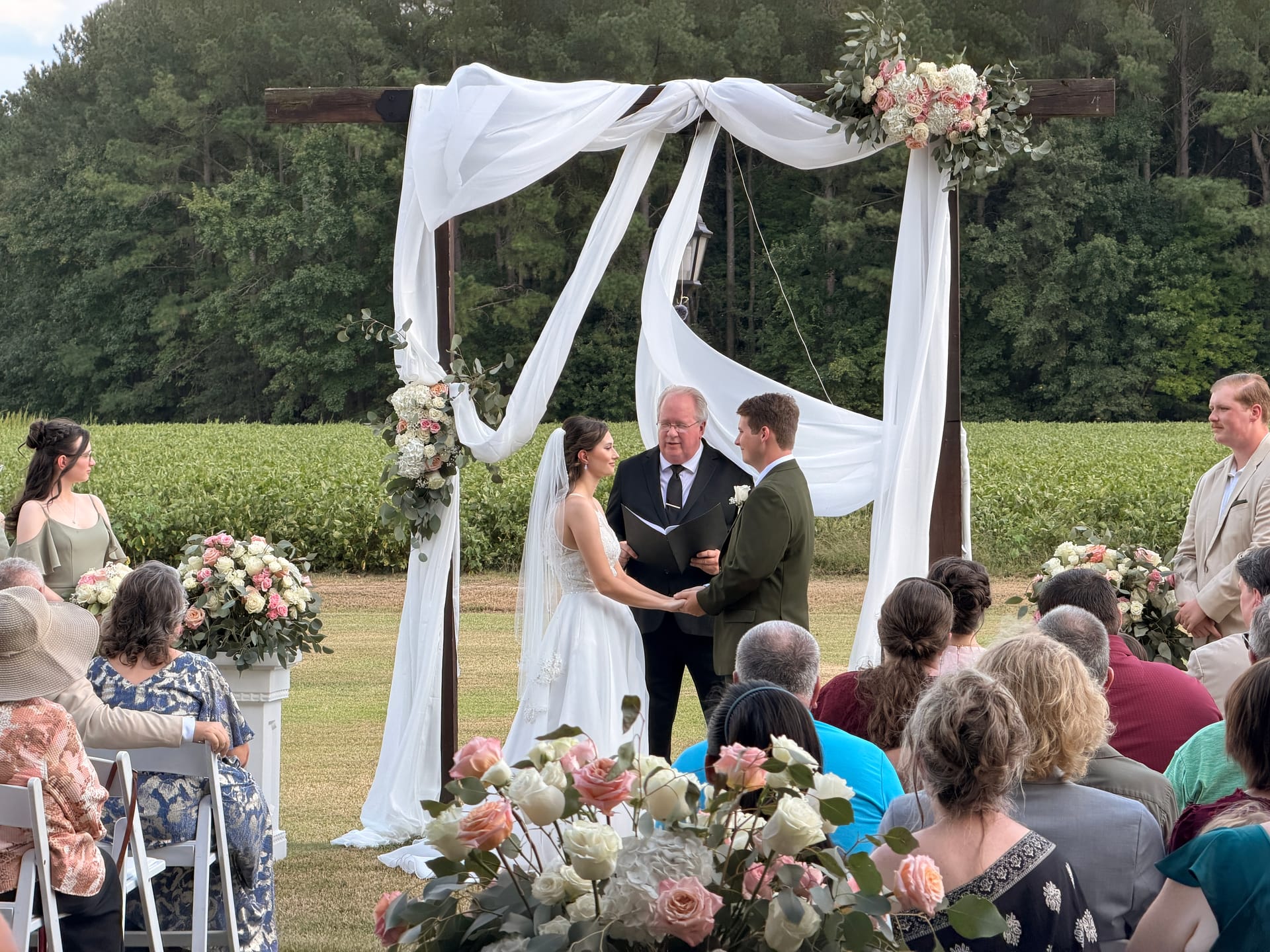 Bride and groom exchange vows beneath a draped wooden arch adorned with blush and white florals on the ceremony lawn at Scarborough House, with green farmland fields stretching behind them.