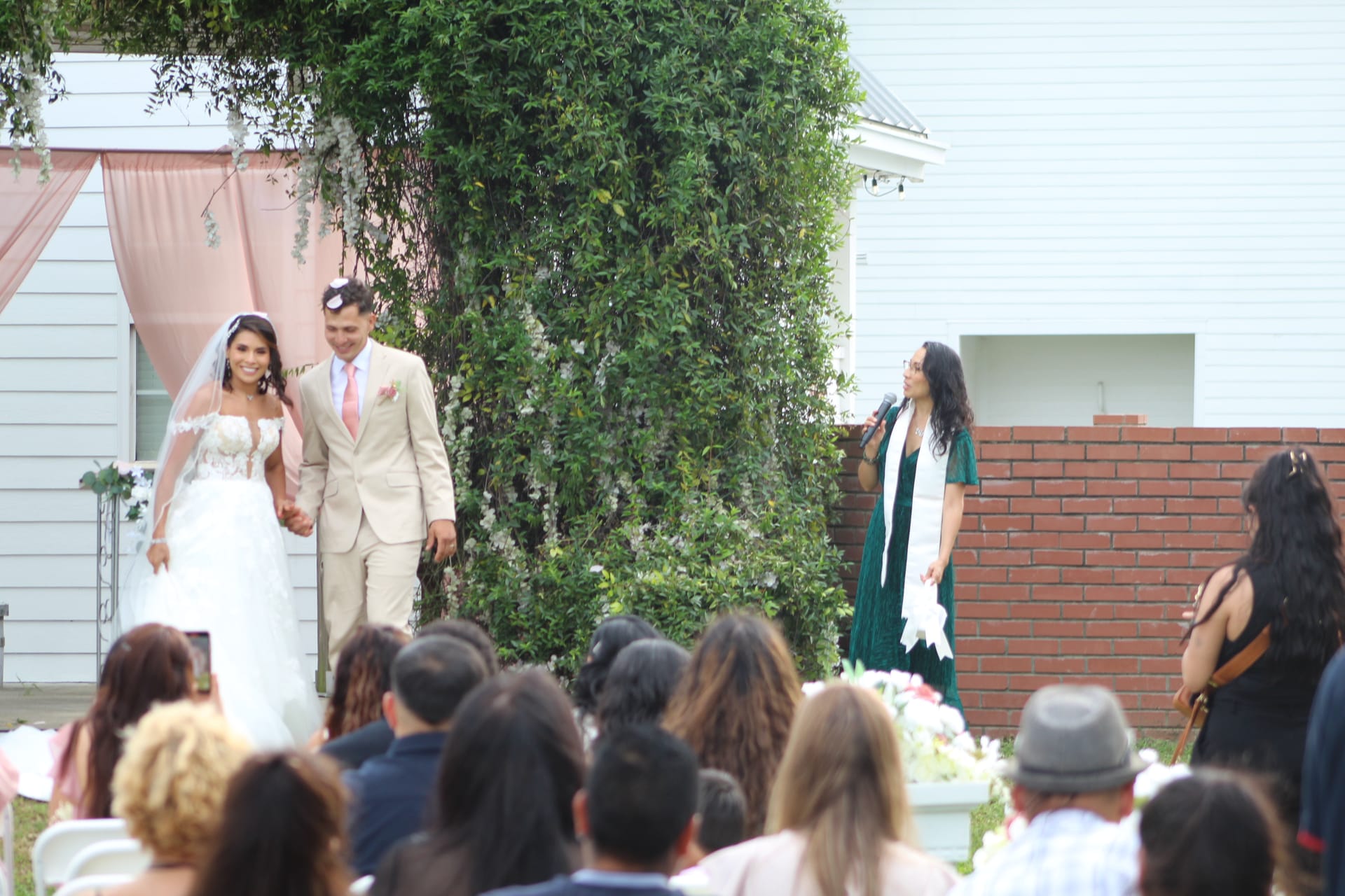 Smiling bride in lace gown and groom in tan suit walk hand-in-hand down the aisle after their outdoor wedding ceremony at Scarborough House as the officiant announces them to seated guests.