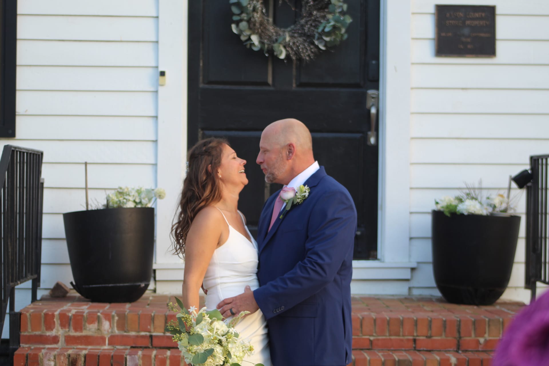 Bride in a white slip gown holding a greenery bouquet laughs joyfully as her groom in a navy suit and pink tie gazes at her on the front steps of the historic 1821 Scarborough House manor.