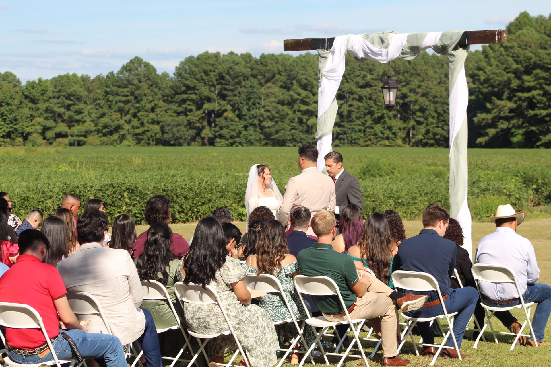 Guests seated in white folding chairs watch a couple exchange vows beneath a rustic wooden arch adorned with white and sage draping at an outdoor farm-field ceremony on a sunny day.