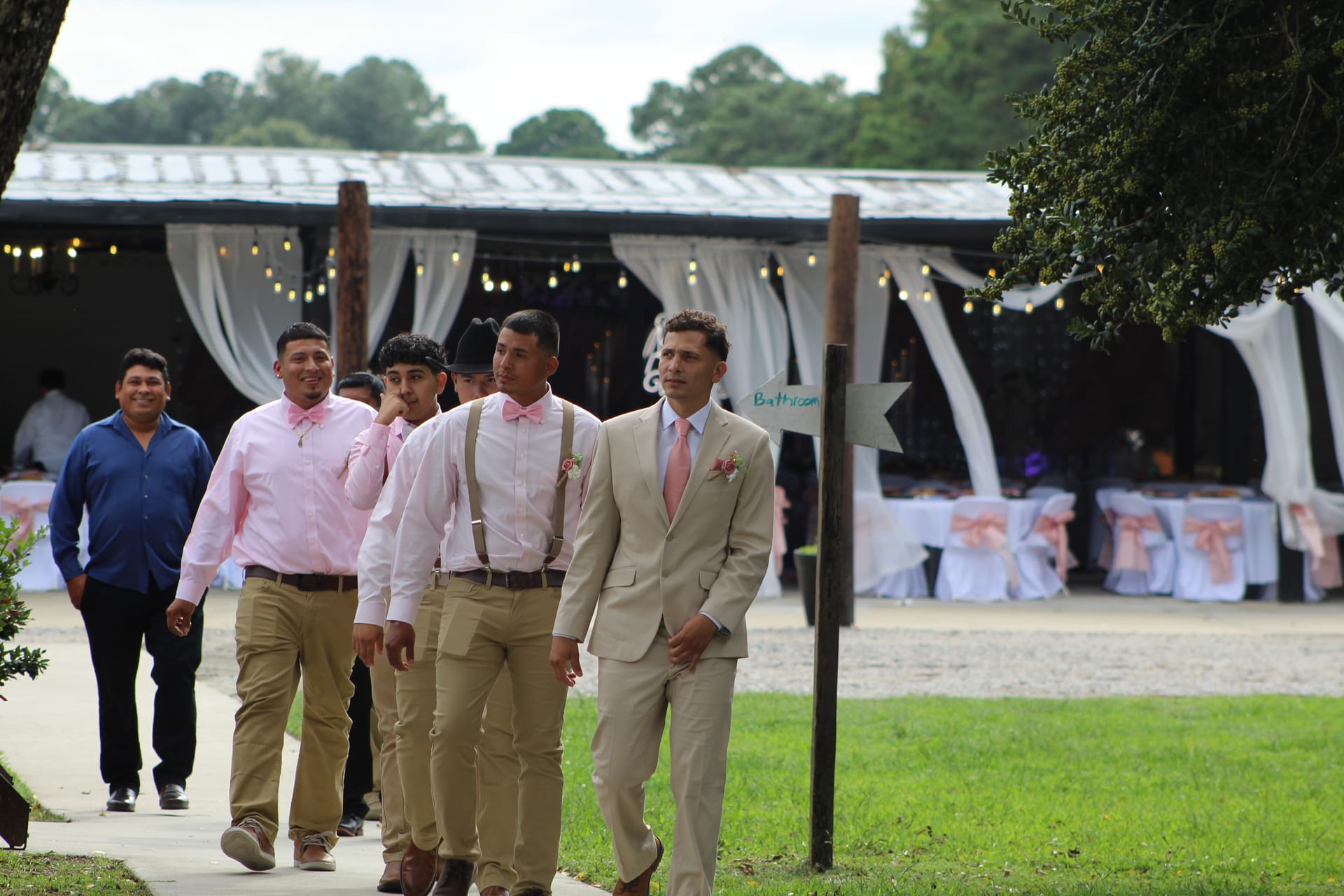 Groom in a tan suit with pink tie leads his groomsmen in matching pink bow ties and suspenders along a garden path toward the decorated reception pavilion at Scarborough House.