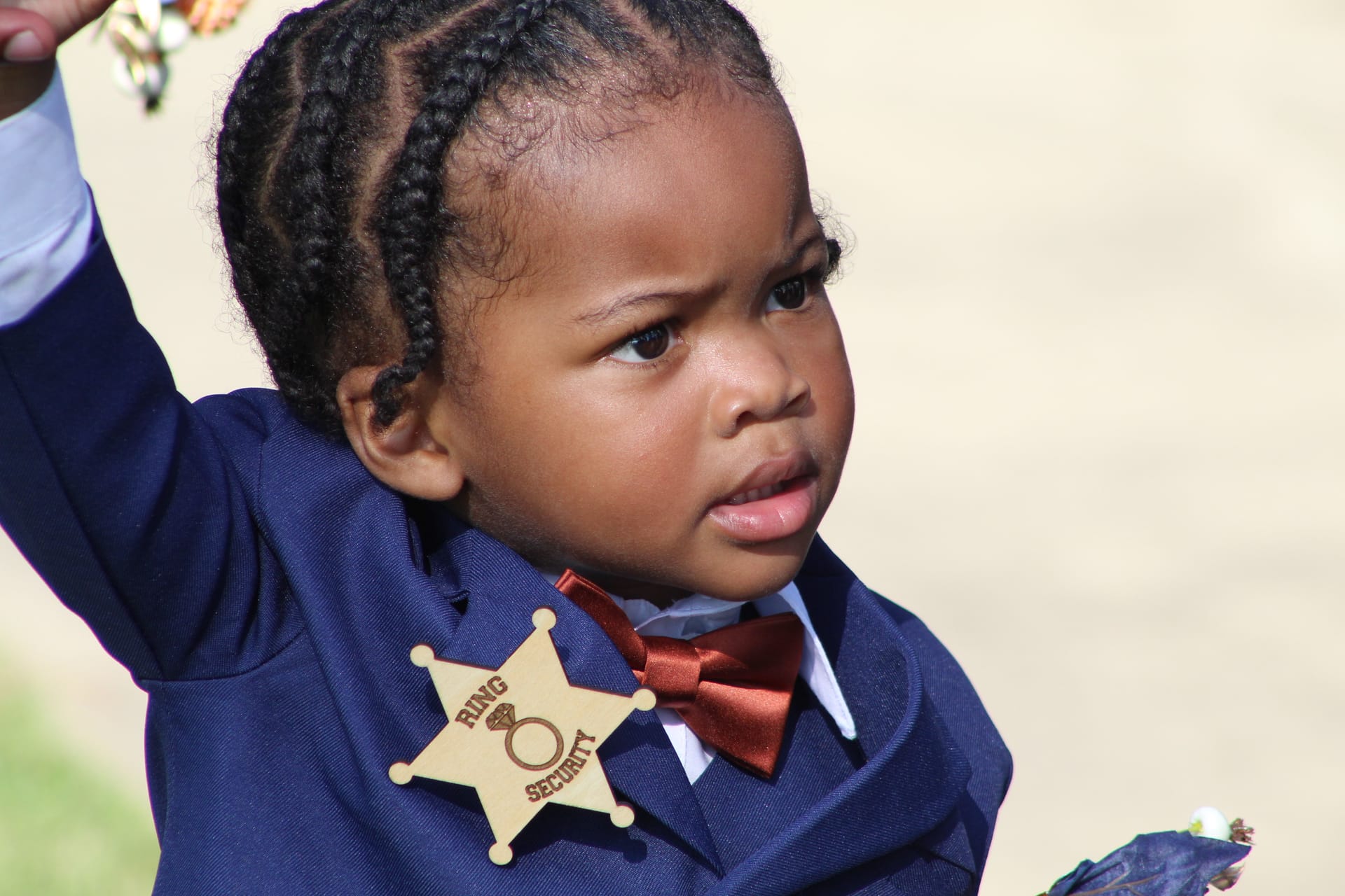 Young ring bearer in a navy suit with a red bow tie and wooden Ring Security sheriff badge raises his arm at an outdoor wedding ceremony.