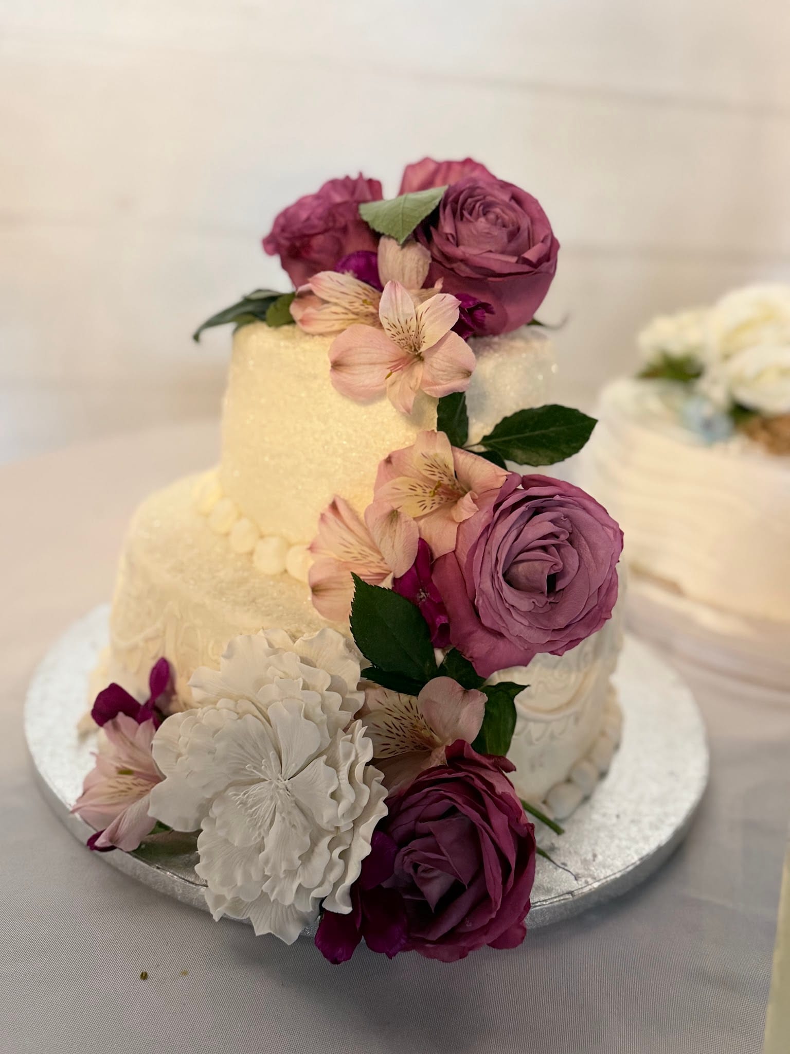 Elegant two-tier white buttercream wedding cake adorned with cascading purple roses, pink alstroemeria, and white hydrangeas on a silver cake board at Scarborough House.