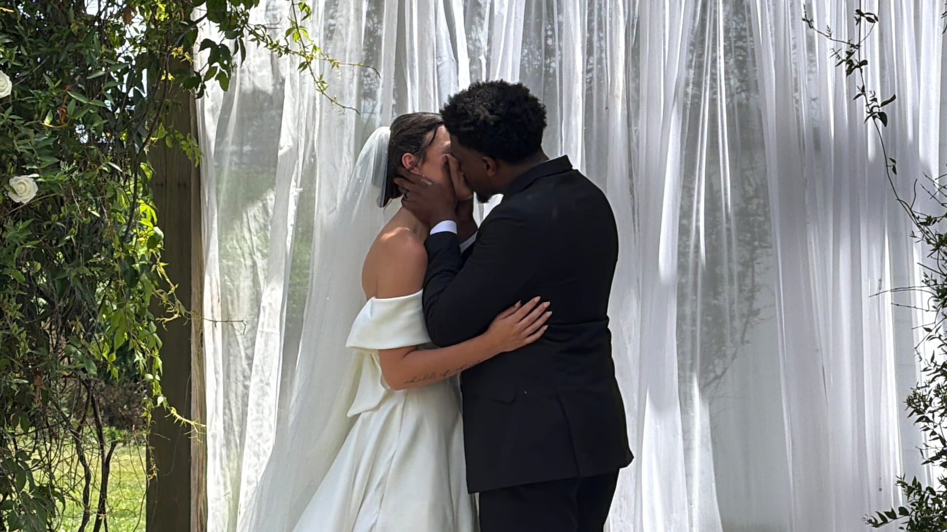Bride in an off-shoulder white gown and veil shares her first kiss with groom in a black suit beneath flowing white drapery at Scarborough House wedding venue.