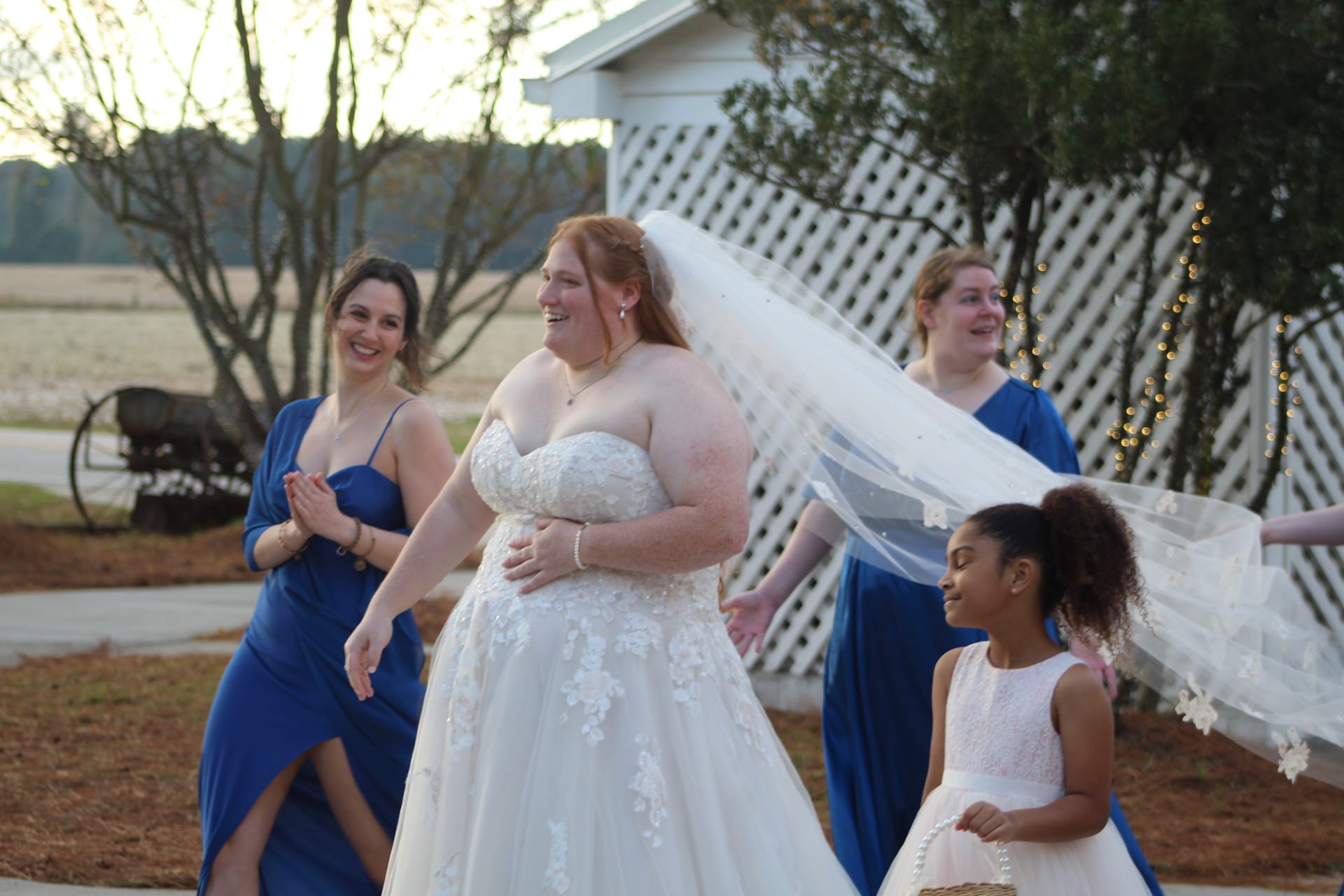 Laughing bride in lace ballgown walks with bridesmaids in royal blue and a flower girl as her veil catches the breeze against the historic Scarborough House farmland backdrop at golden hour.