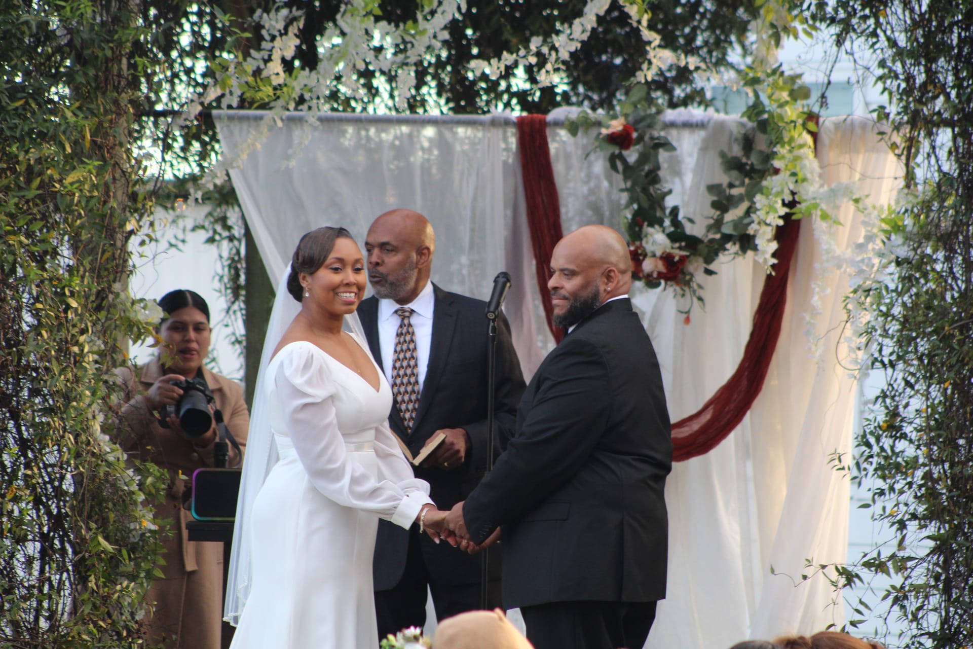 Bride in a white long-sleeve gown smiles joyfully while holding hands with her groom in a black tuxedo during their outdoor wedding ceremony beneath a draped floral arch at Scarborough House.