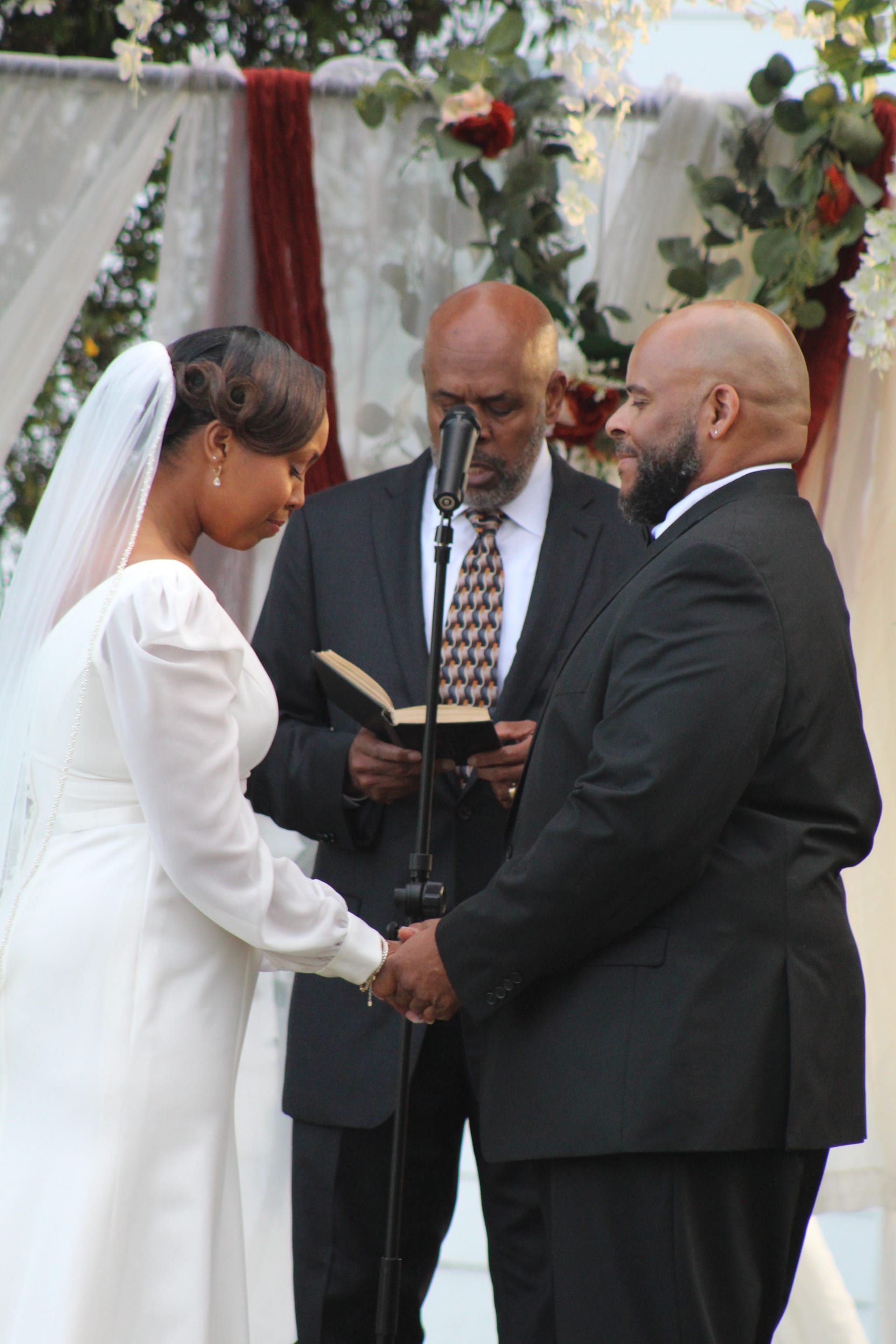 Bride in a long white gown and groom in a black suit hold hands during their outdoor wedding ceremony while an officiant reads from a book beneath a floral and draped arch.