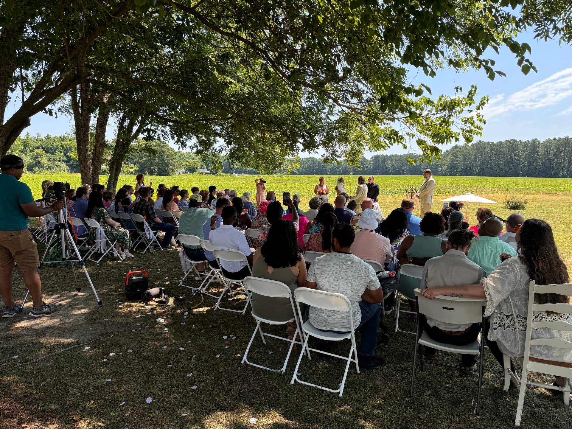 Outdoor wedding ceremony at Scarborough House with guests seated on white folding chairs under large oak trees overlooking a lush green farmland field on a sunny summer day.