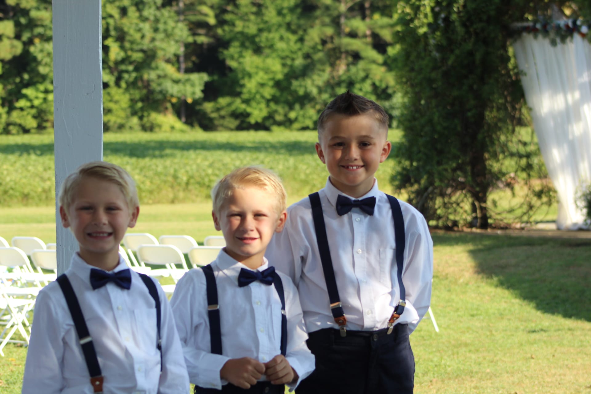 Three young ring bearers dressed in white shirts, navy bow ties, and suspenders smile together on the sunlit ceremony lawn at Scarborough House wedding venue.