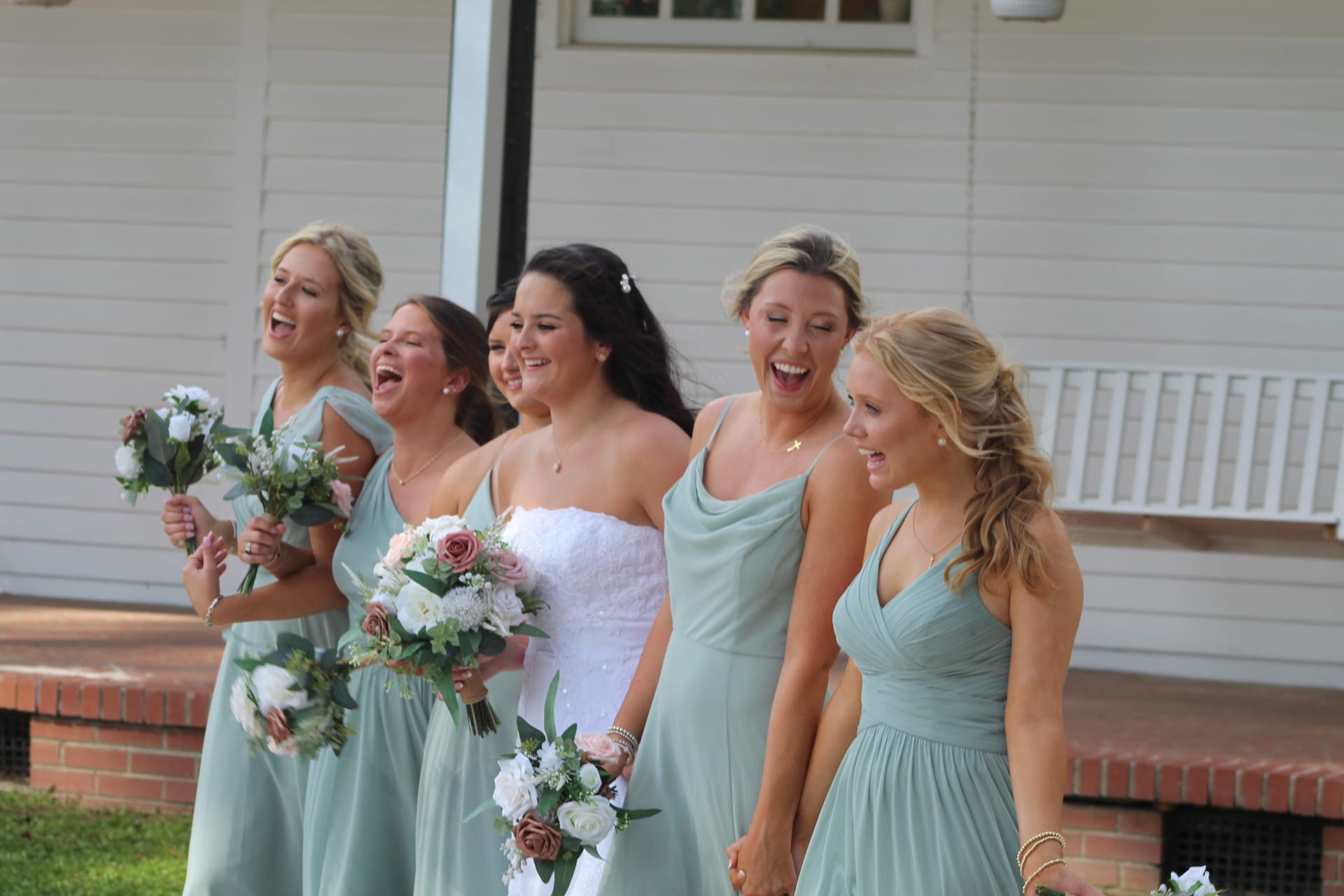 Bride in white gown laughing with five bridesmaids in sage green dresses holding floral bouquets outside the historic Scarborough House manor.