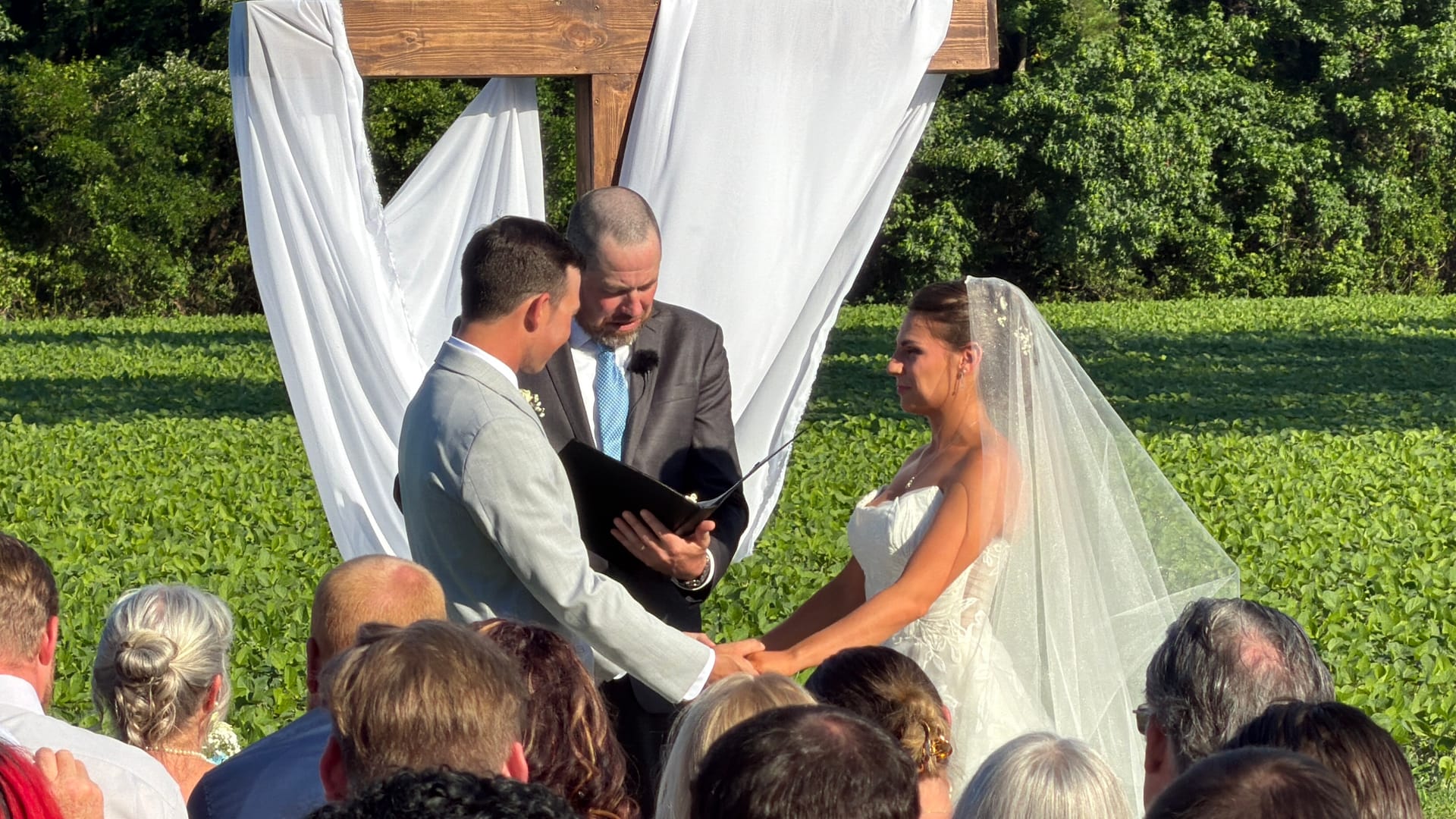 Bride and groom hold hands during their outdoor wedding ceremony beneath a draped wooden cross arbor with green farmland fields in the background.