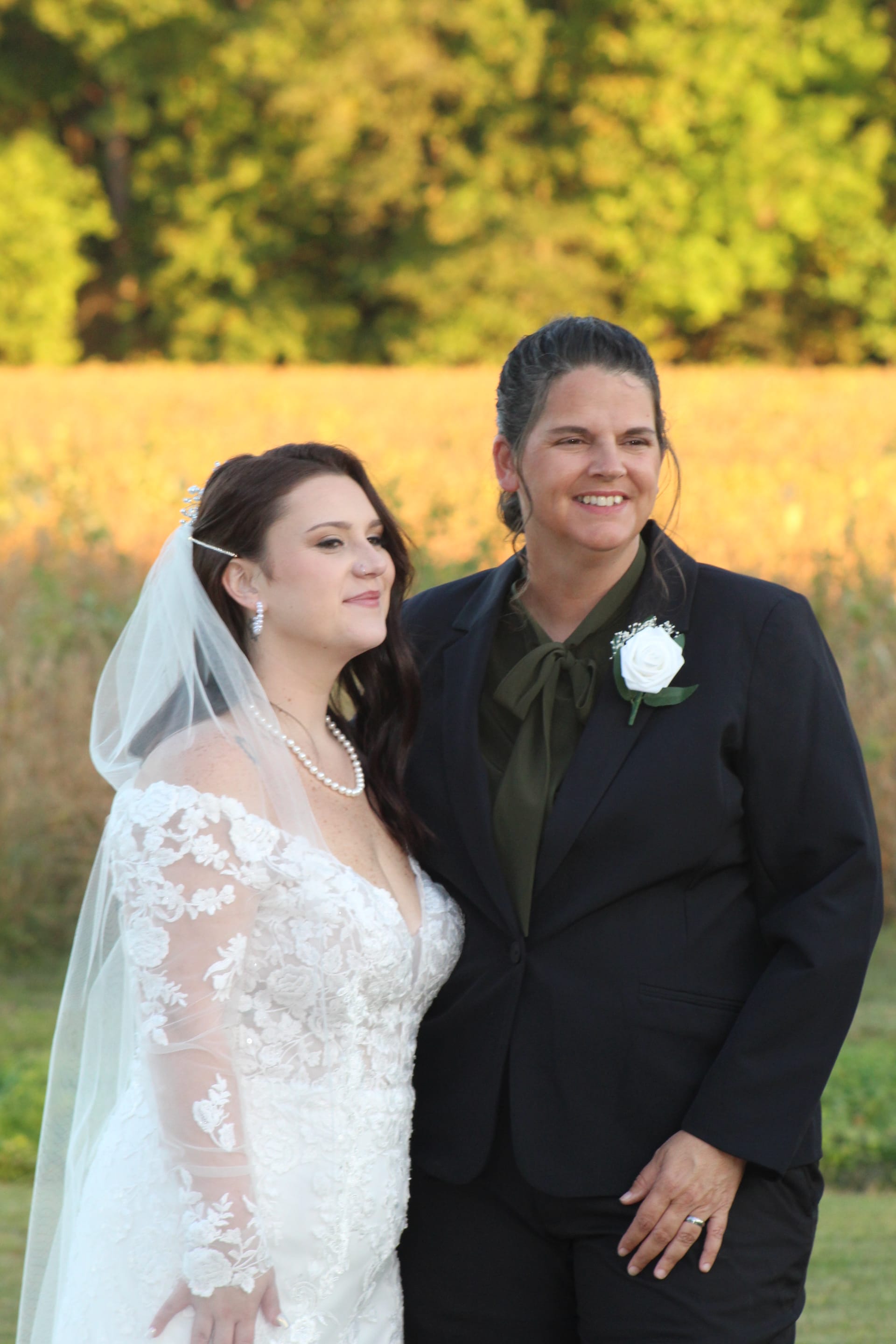 Two brides pose together at golden hour in front of a sunlit farmland field, one in a lace wedding gown with veil and the other in a navy suit with white boutonniere, at Scarborough House estate.